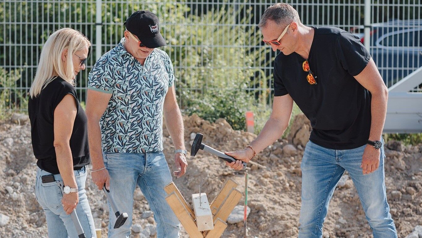 Three people are standing around a wooden structure and working with hammers to secure an object. Background with trees and fence.