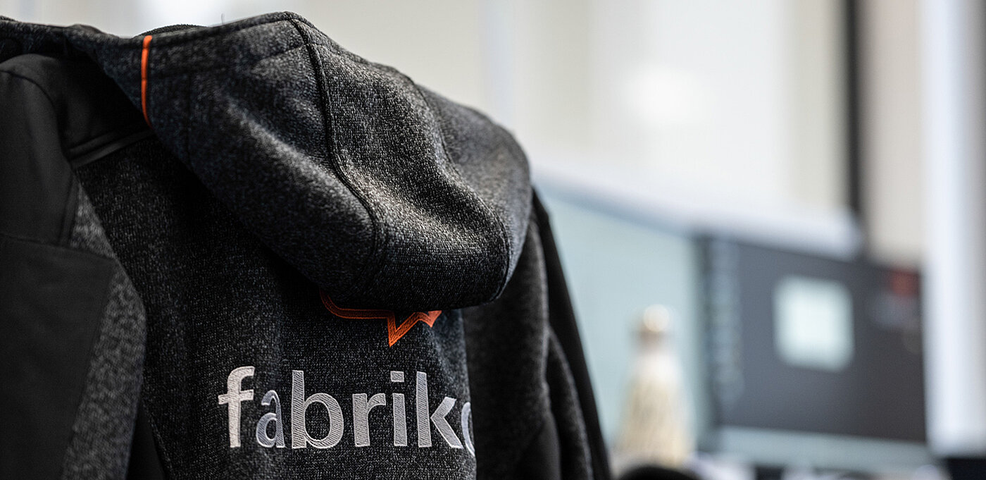 Hoodie mit Kapuze und dem Schriftzug "fabrik" in einer modernen Büroumgebung.
