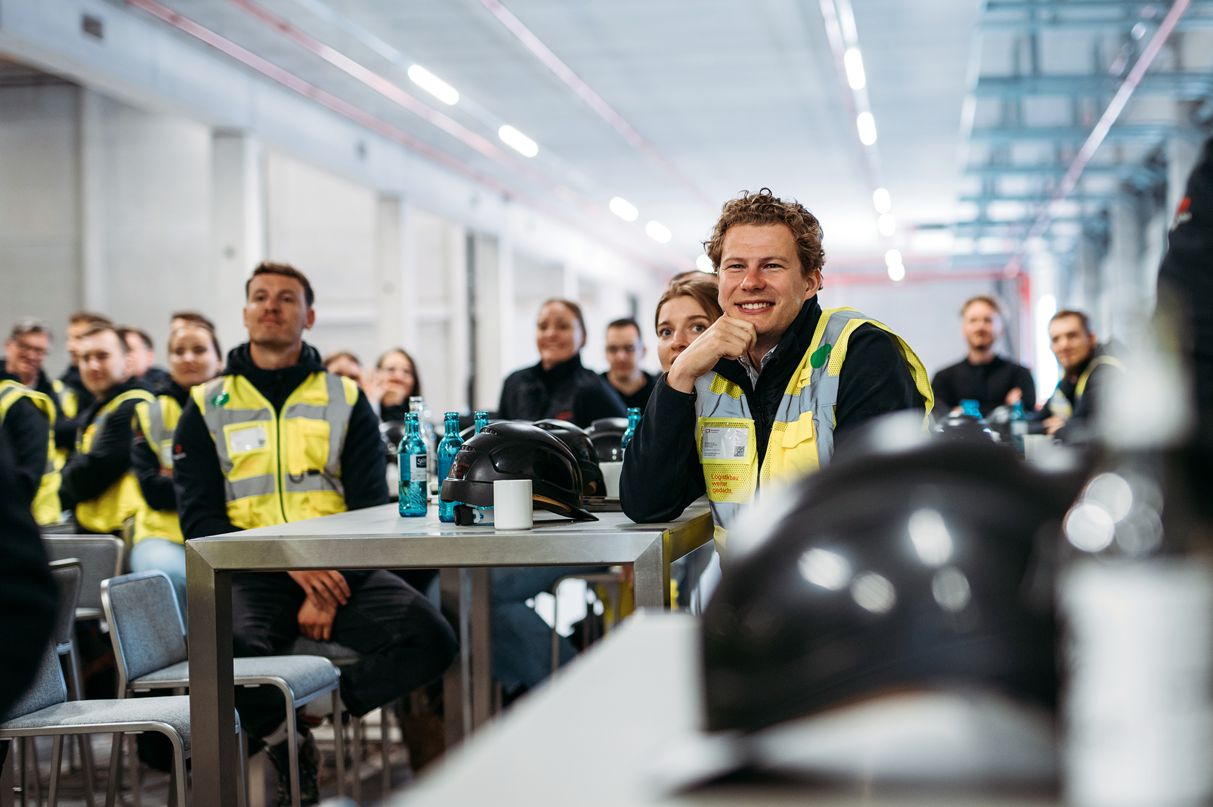 fabrikon Mitarbeitende in Warnwesten verfolgen aufmerksam einen Vortrag in einer Industriehalle; Schutzhelme und Wasserflaschen stehen auf den Tischen.