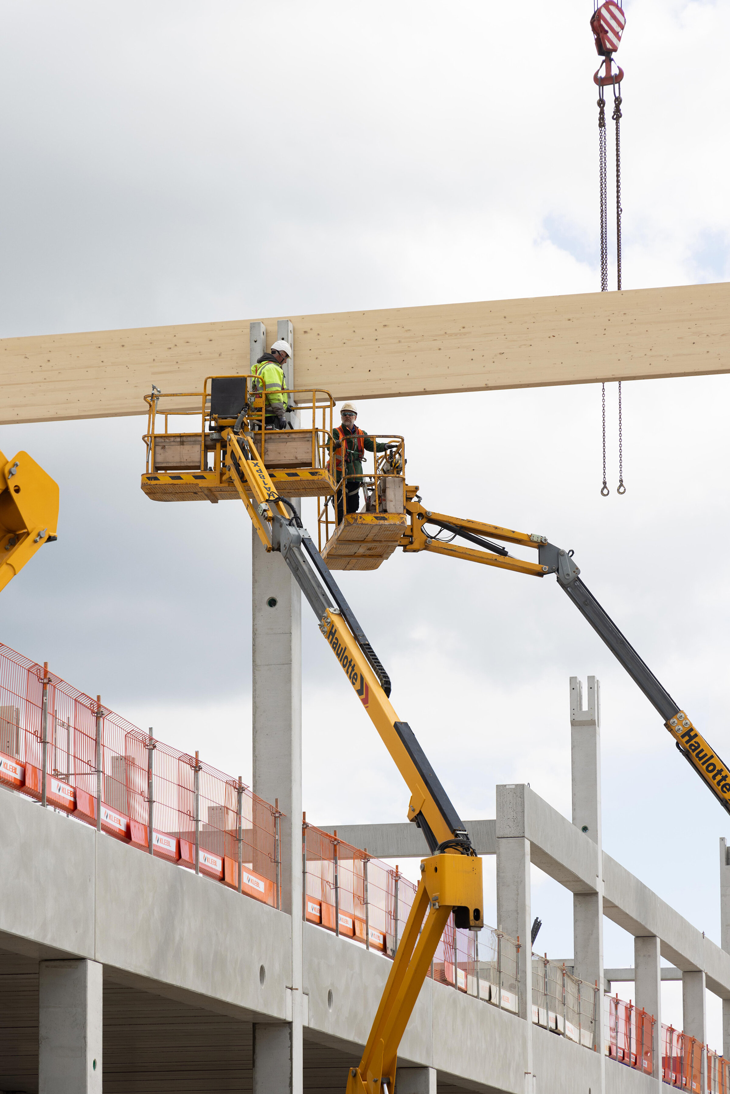 Zwei Bauarbeiter arbeiten auf einer Hebebühne an einem Holzträger eines Baustellengebäudes.