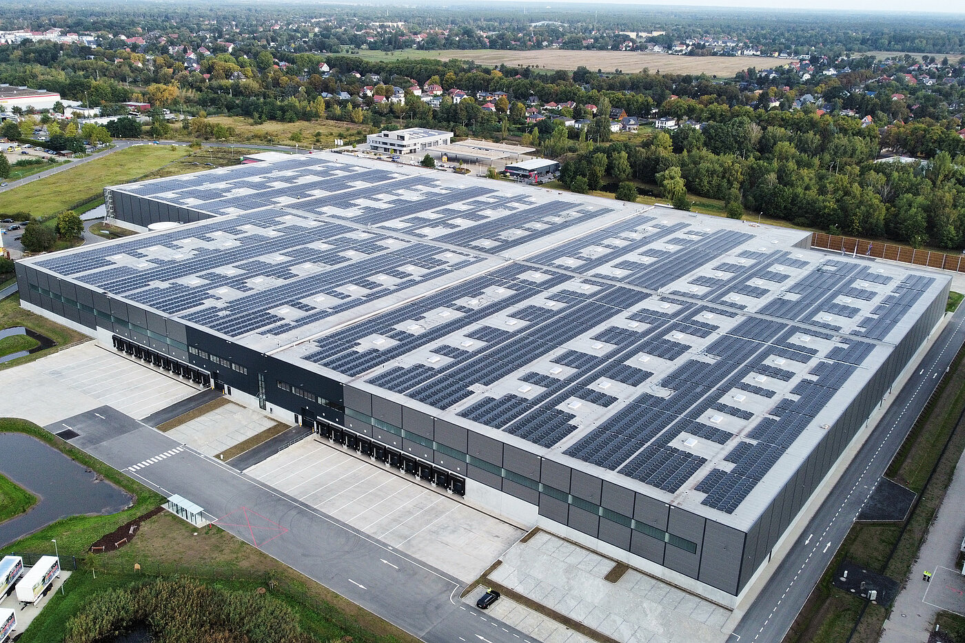 Großes Lagergebäude mit Solarpanelen auf dem Dach, umgeben von einer grünen Landschaft und weiteren Gebäuden.
