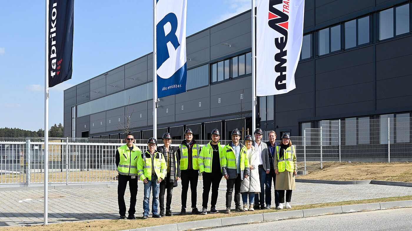 Die Projektbeteiligten am Logistikbau stehen mit Warnwesten zur Schlüsselübergabe vor dem Eingangsbereich an der Nordfassade nach erfolgreicher Übergabe