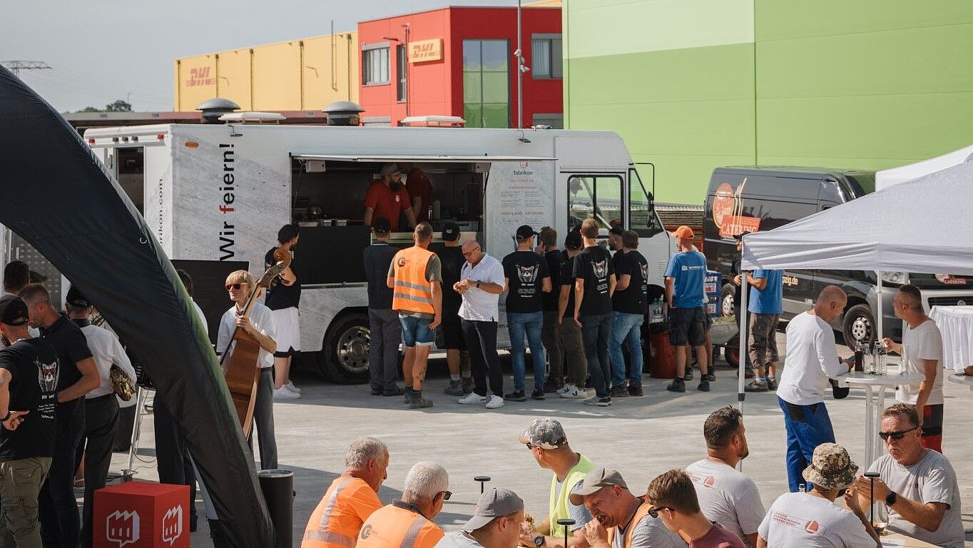 People are standing in front of a food truck on a site, while others are eating and celebrating at tables.