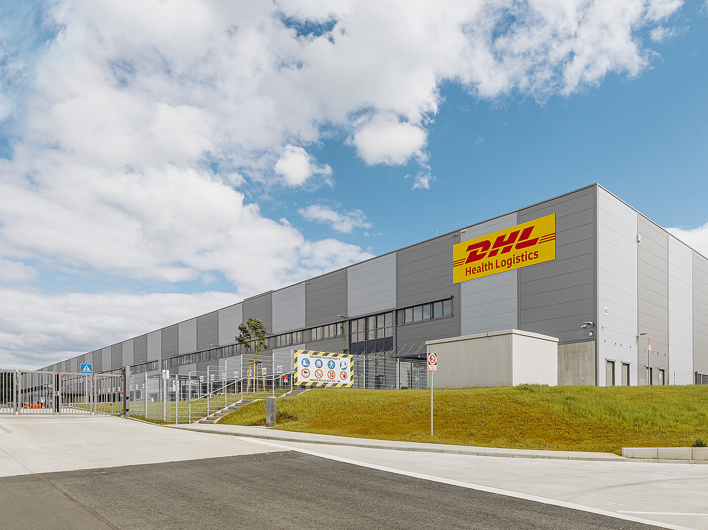 Großes, modernes Lagergebäude von DHL Health Logistics unter einem bewölkten Himmel.