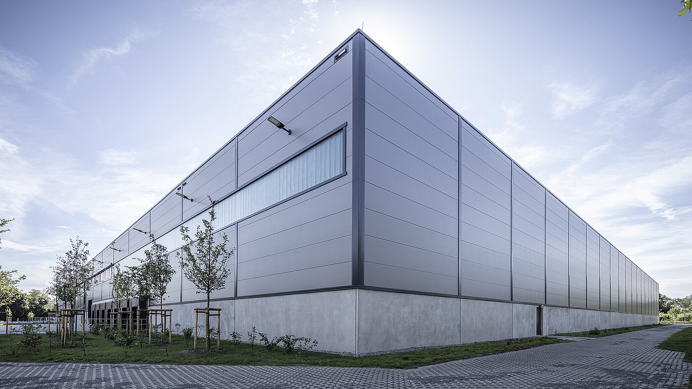 Moderne Lagerhalle mit grauer Fassade, umgeben von Bäumen und Gras, im Hintergrund ein klarer Himmel.
