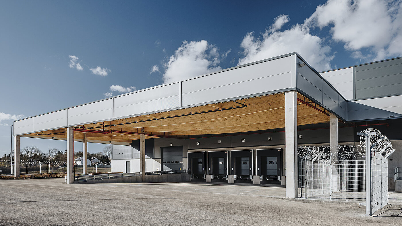 Moderne Lagerhalle mit Überdachung, Laderampen und Sicherheitszaun bei klarem Himmel.