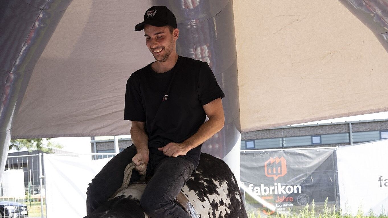 Young man on a mechanical bull, smiling, in a tent with a colorful floor and a banner in the background.