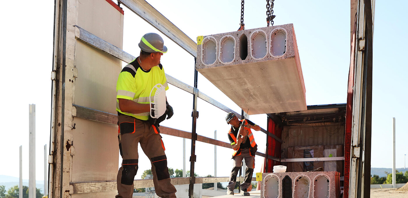Zwei Bauarbeiter in Warnkleidung entladen Betonblöcke von einem Lkw unter sonnigem Himmel.