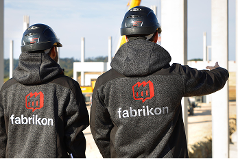 Zwei Personen mit Helmen und Jacken mit dem Logo "fabrikon" stehen auf einer Baustelle und schauen in die Ferne.