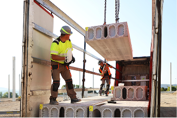 Zwei Bauarbeiter entladen Betonblöcke von einem Lkw mit einem Kran bei sonnigem Wetter.