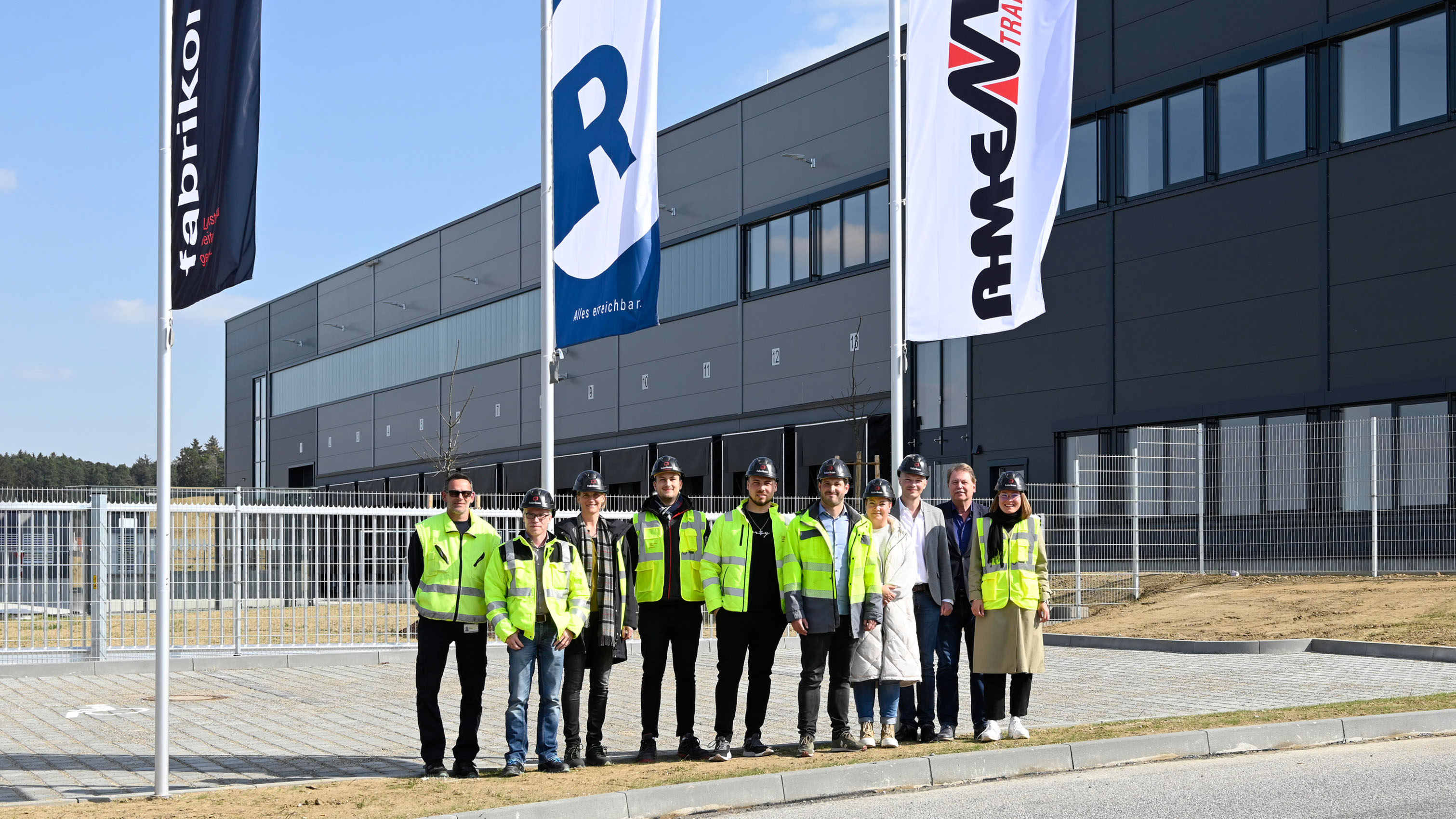 Die Projektbeteiligten am Logistikbau stehen mit Warnwesten zur Schlüsselübergabe vor dem Eingangsbereich an der Nordfassade nach erfolgreicher Übergabe