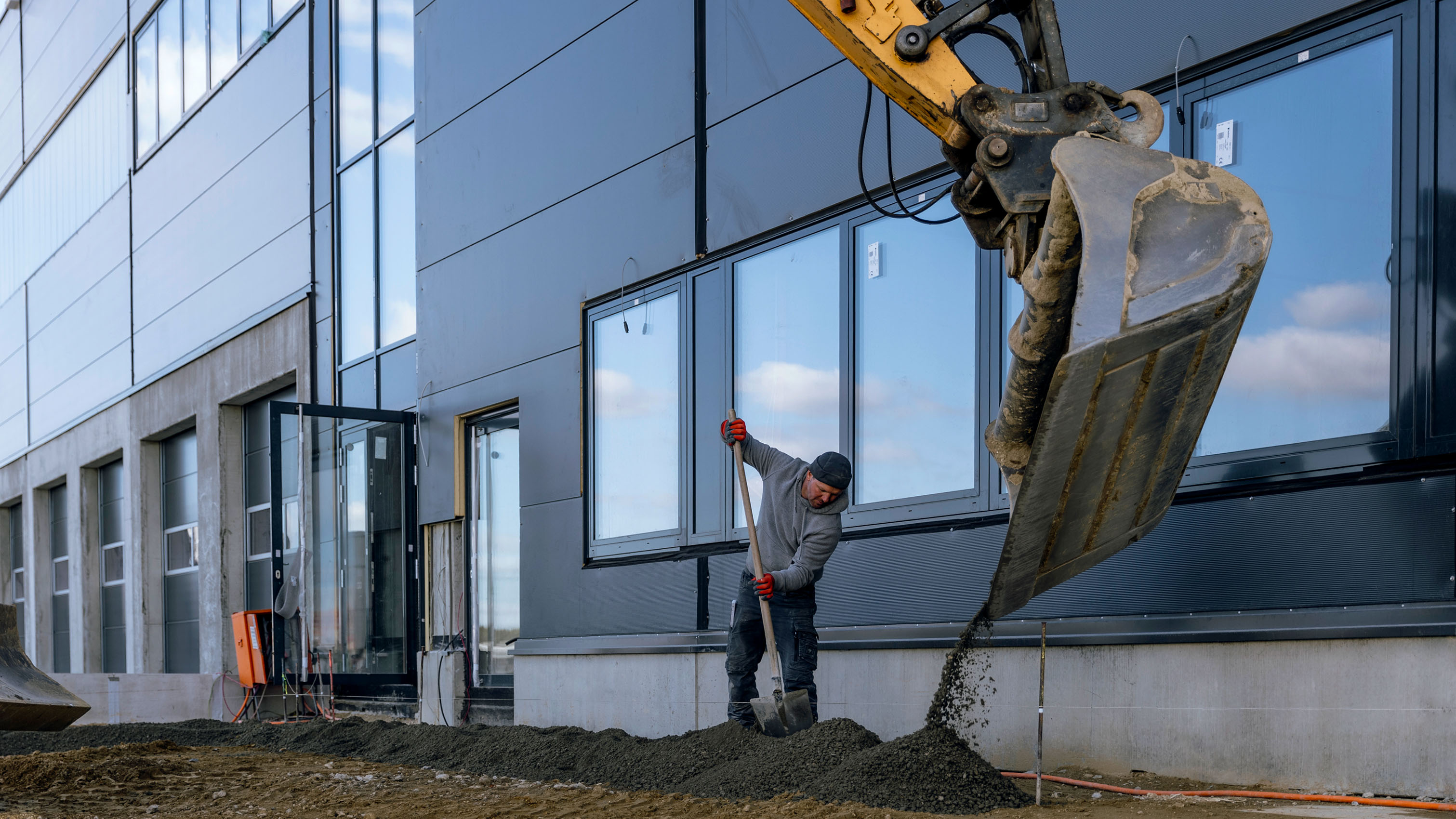 Riesige Baggerschaufel und Bauarbeiter mir Schaufel vor Logistikbau-Fassade