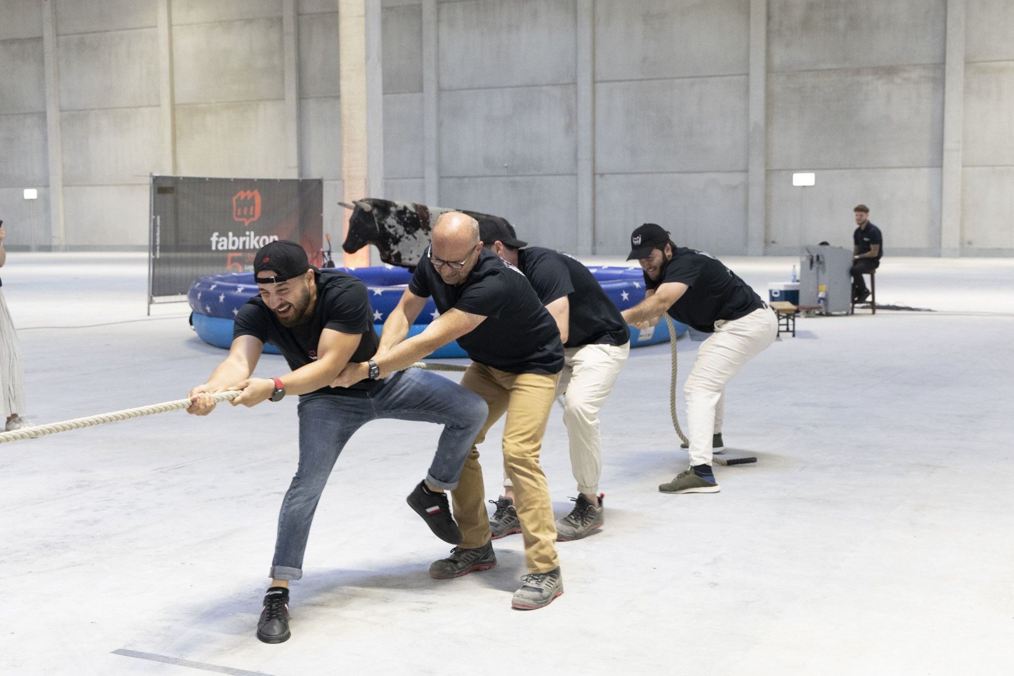 Four men pull on a rope in a large hall, while an attraction is visible in the background.