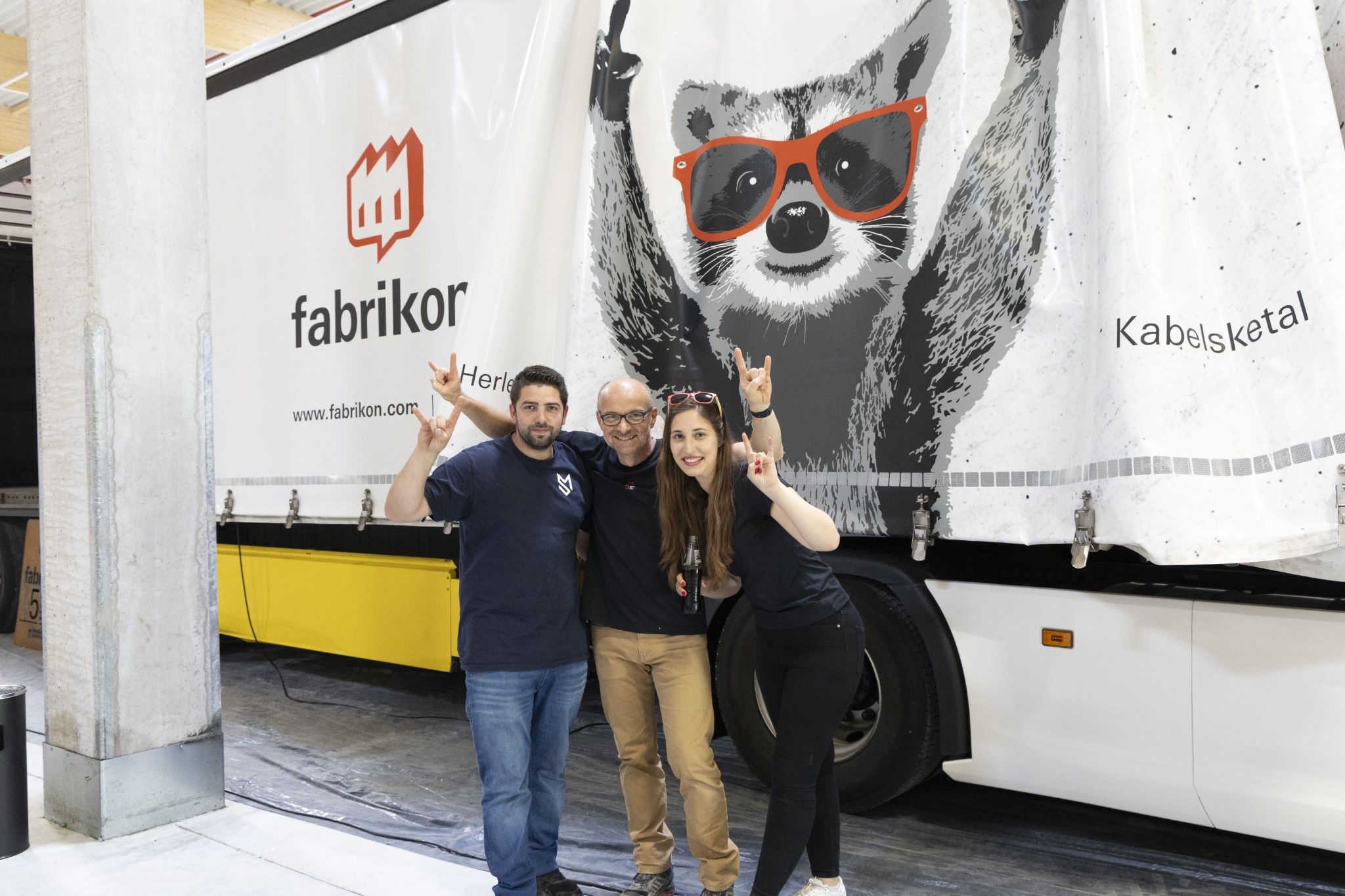 Three people pose in front of a truck with an animal motif and the lettering "fabrikor." They all make the peace sign.
