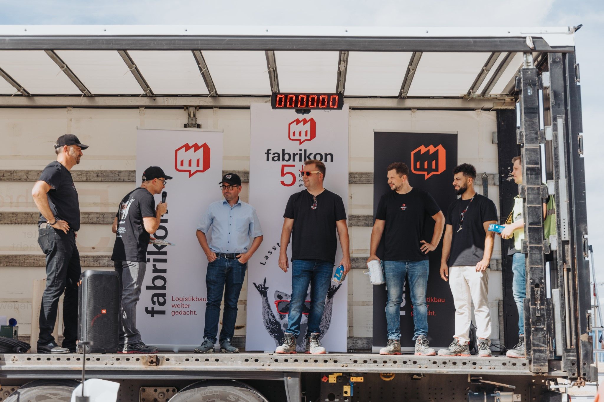 Four men stand on a stage while one speaks; in the background are banners of Fabrikon.