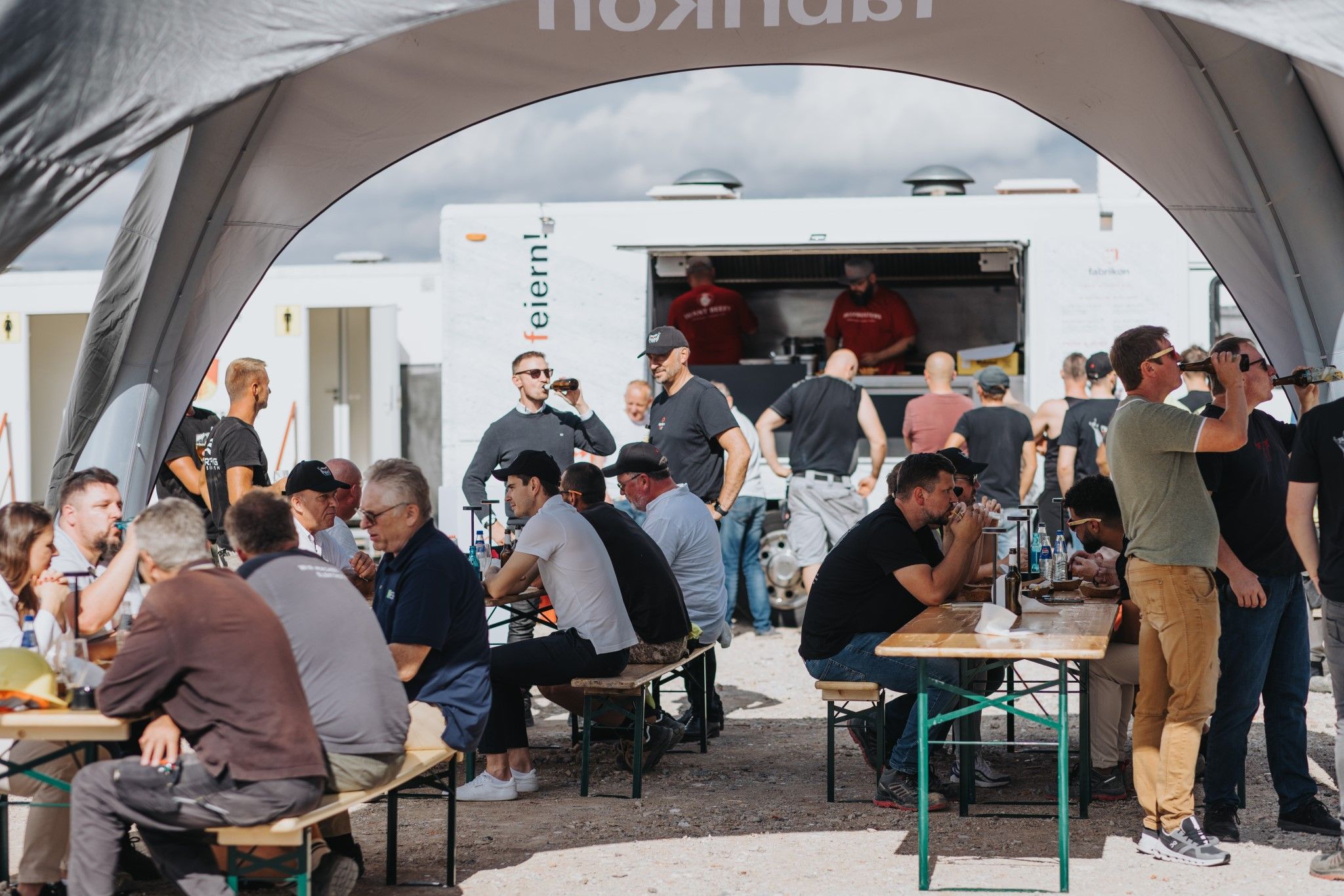 People are sitting and standing under a tent, eating and drinking, with a food truck in the background.