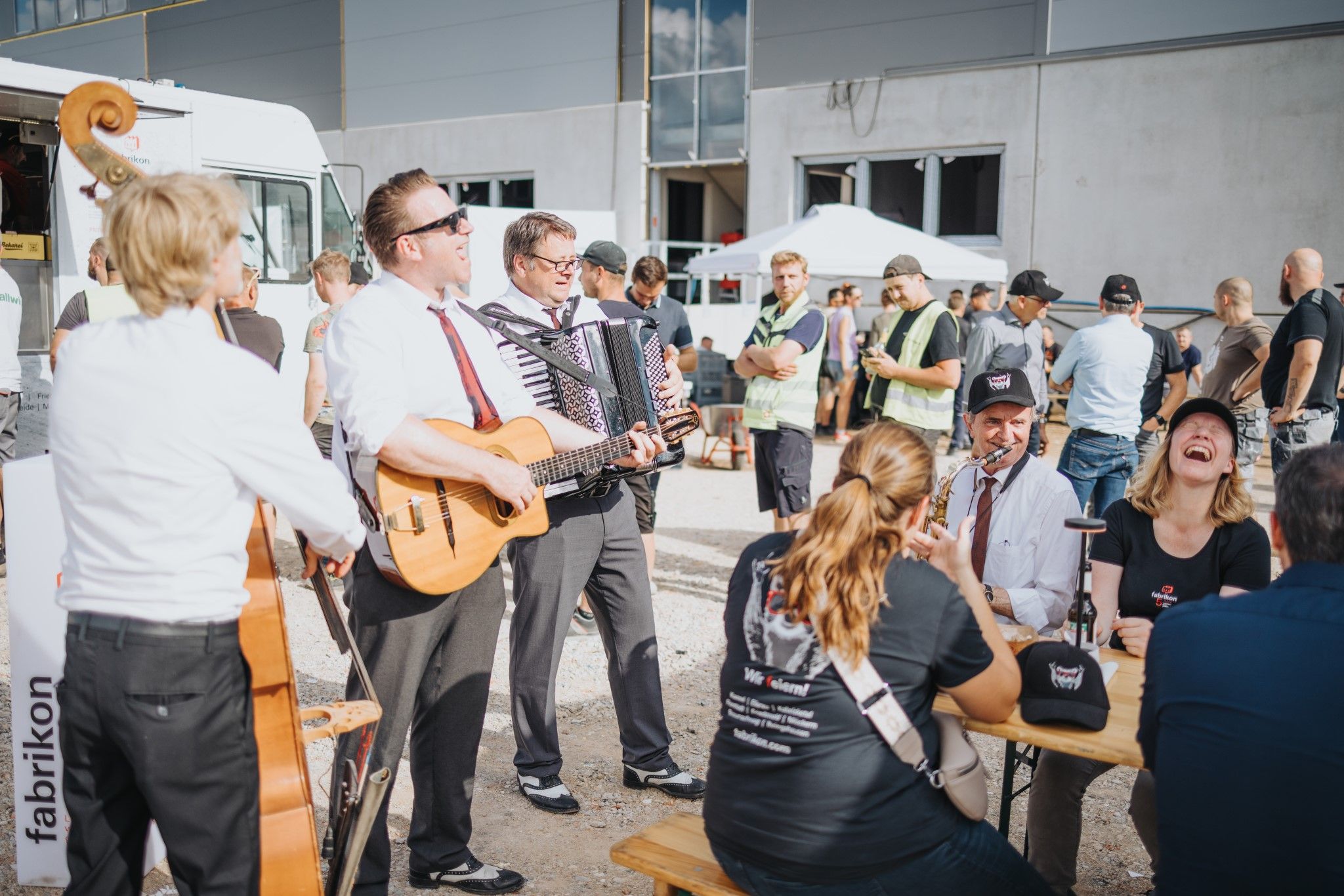 A band plays at an event, people laugh and enjoy the atmosphere, with stands and spectators in the background.
