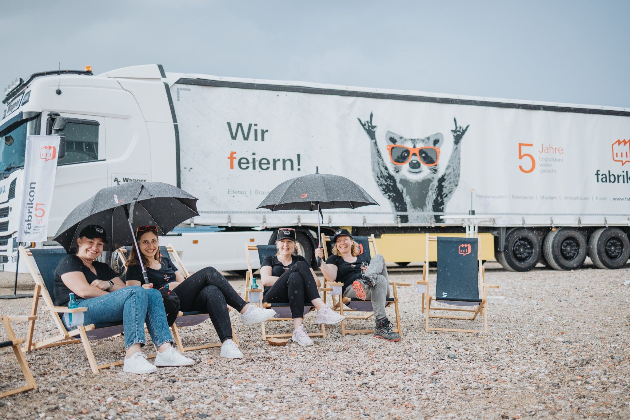 Four people with umbrellas are sitting relaxed on folding chairs in front of a truck with the words "We're celebrating!" printed on it.
