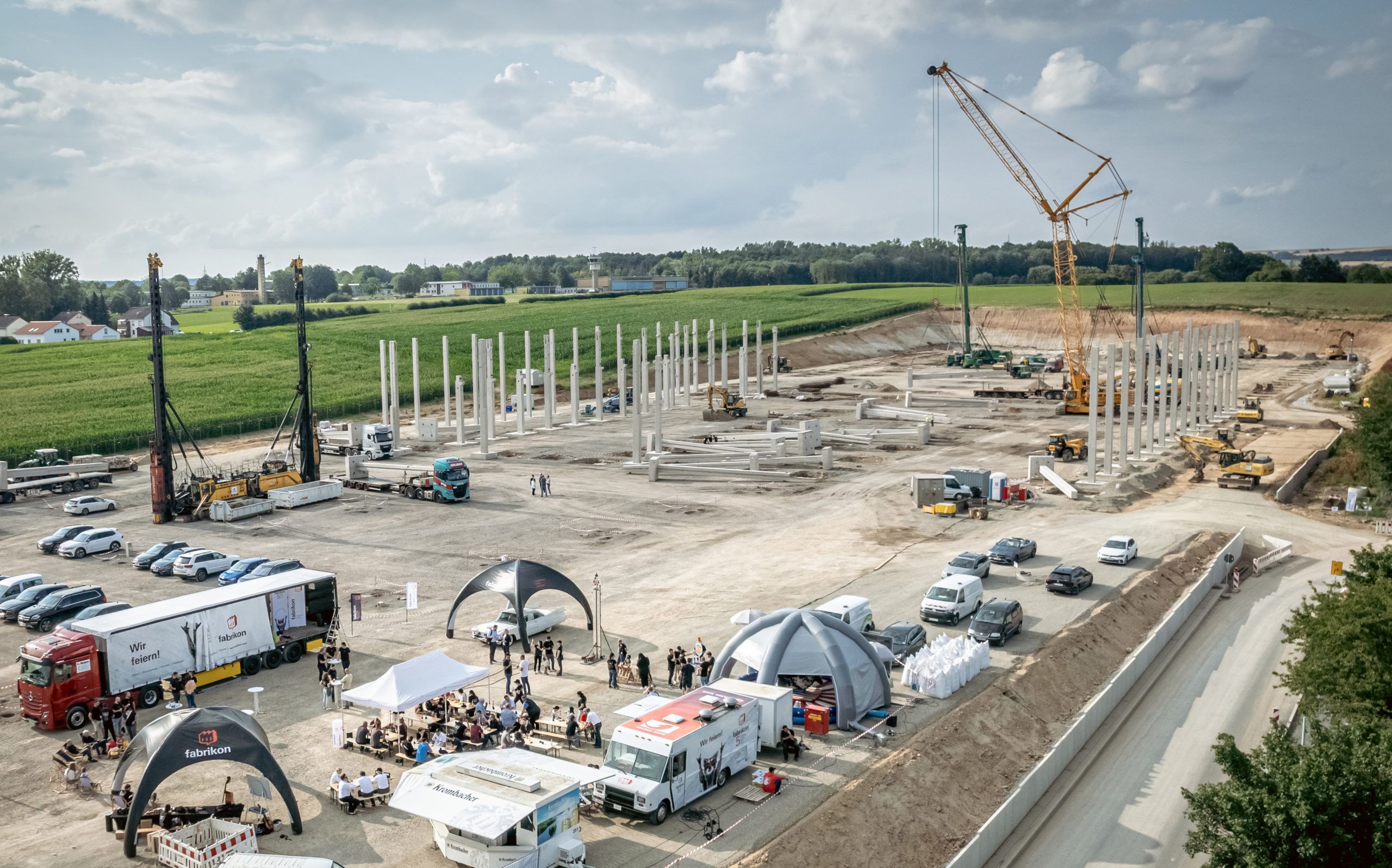 Construction site with cranes, construction machinery, and several work vehicles, concrete pillars, and a party for construction workers.