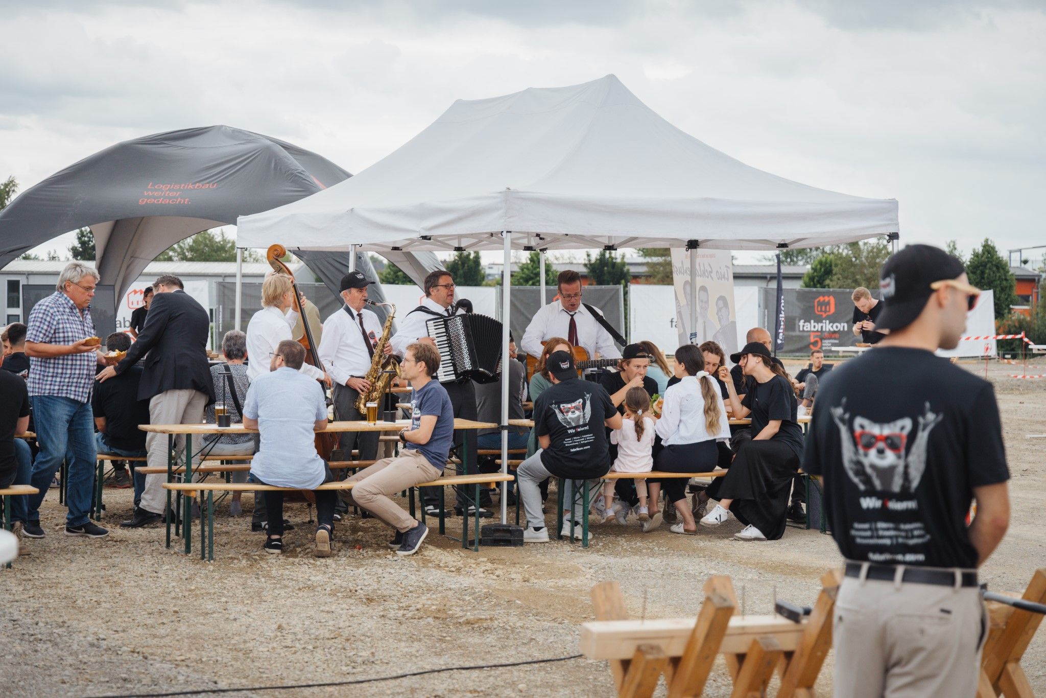 People are sitting in a convivial gathering under pavilions while a band plays live music.