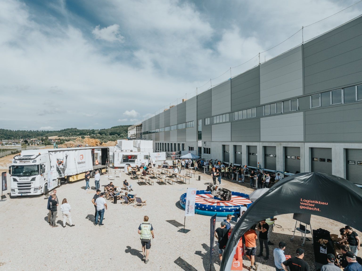 Crowd in front of a warehouse with vehicles, tables, and outdoor events in nice weather.