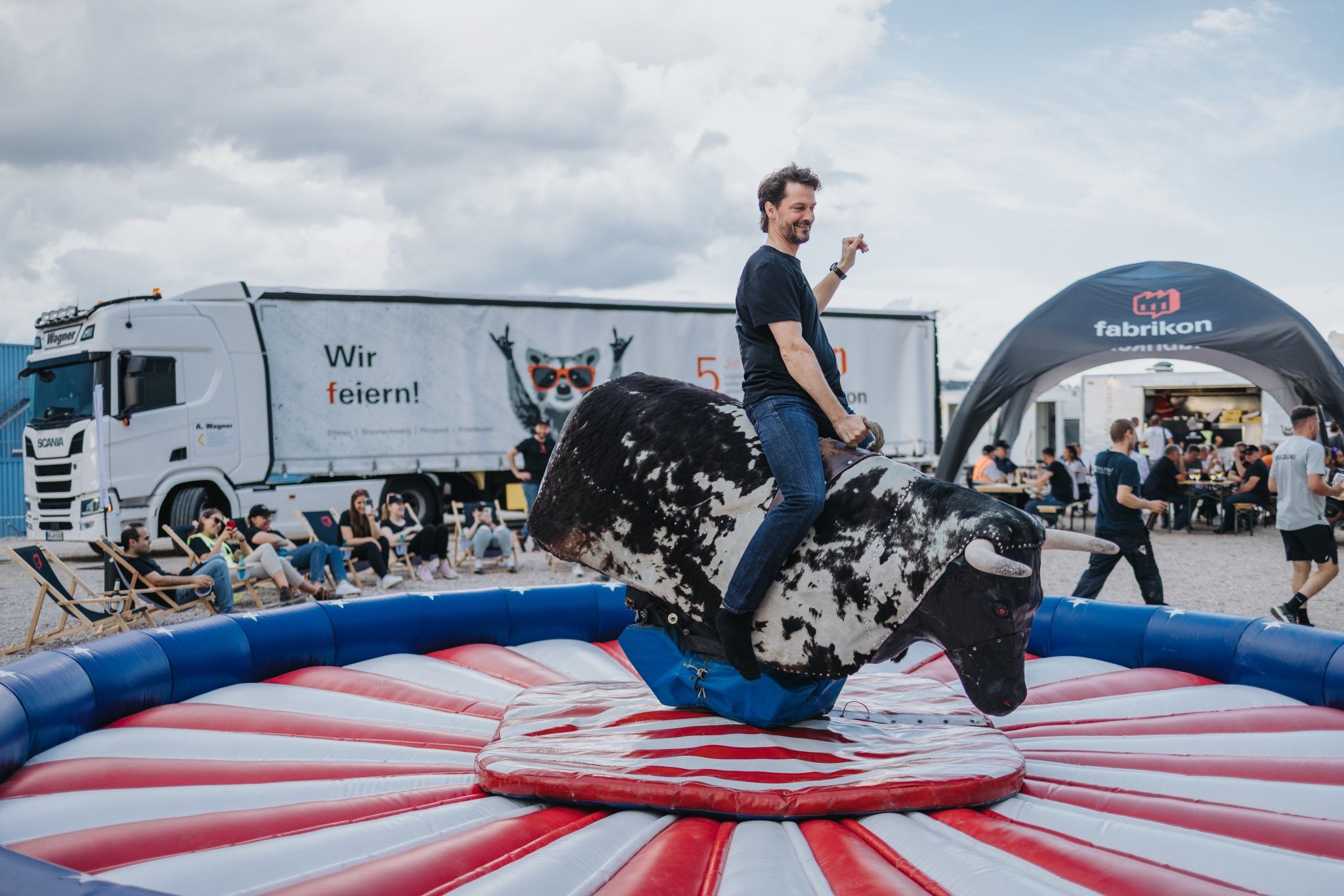 A person is riding a mechanical bull. In the background, spectators and a truck with advertising can be seen.