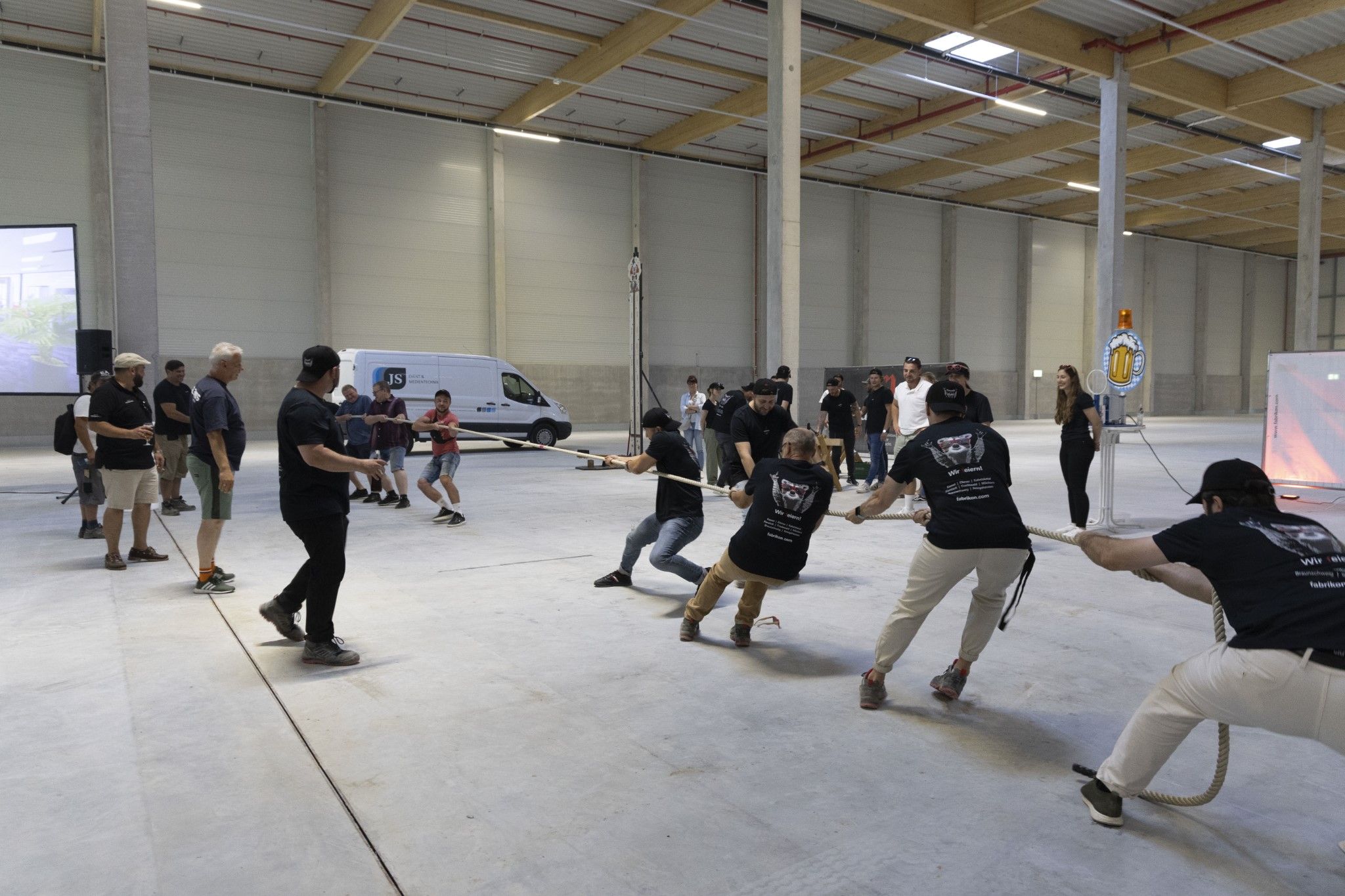 Groups of men pull on a rope during a competition in a large hall. Spectators watch the event.