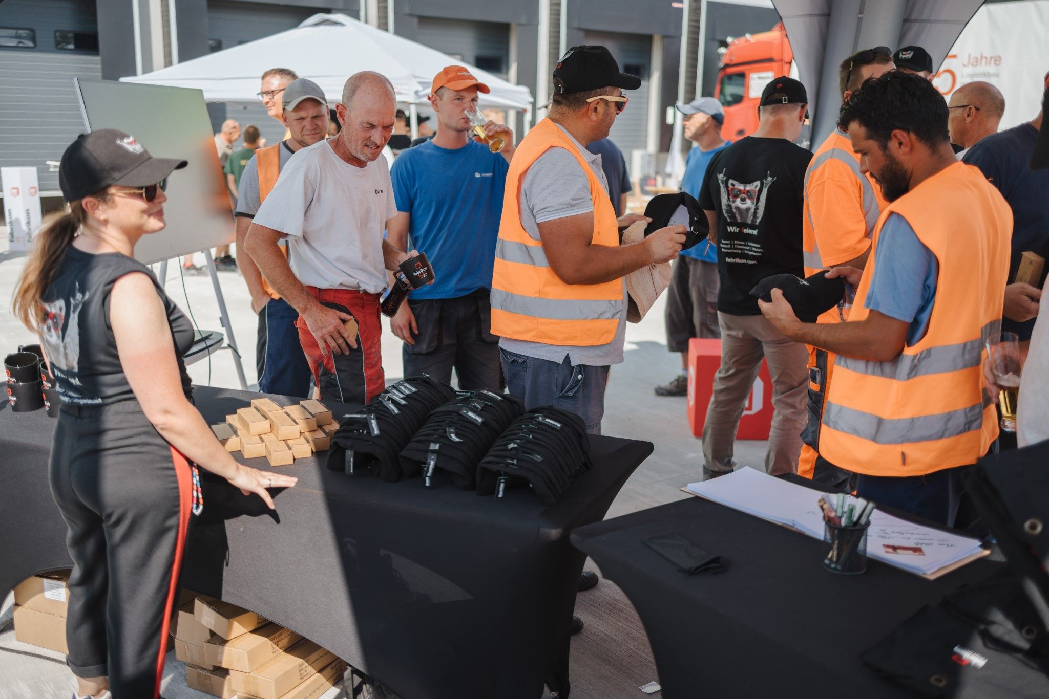 Group of people in work clothes interacting at a stand with hats and promotional materials. Sunny day.