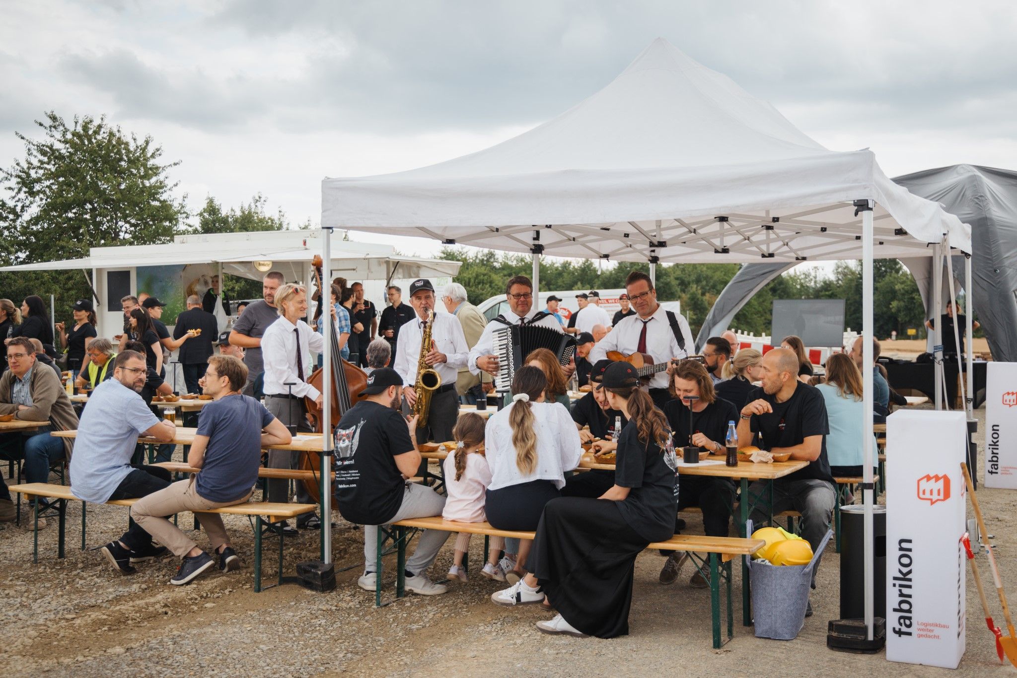 A crowd of people are sitting under a tent while musicians entertain the event with live music. Food and drinks are available.