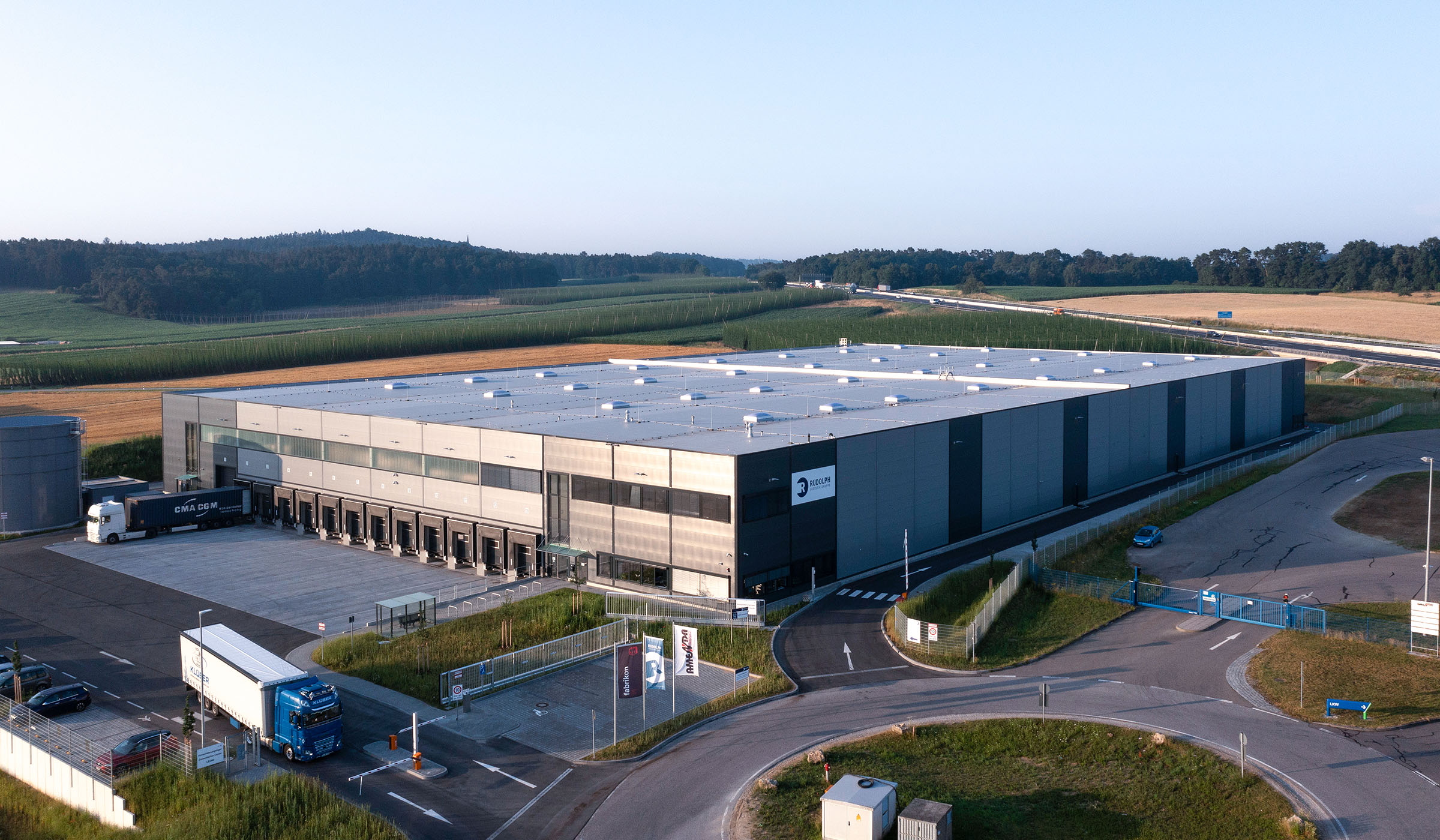 Modernes Lagergebäude mit Lkw-Anlieferungen, umgeben von Feldern und Hügeln, unter klarem Himmel.