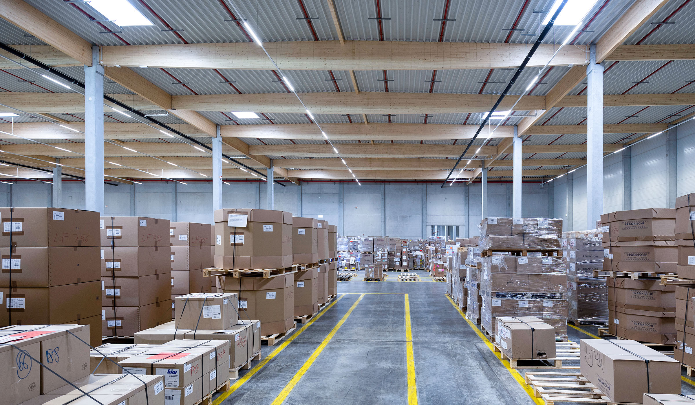 Lagerhalle mit gestapelten Kartons, hell beleuchtet, klare Wege zwischen Regalen, effiziente Anordnung für Lagerung.