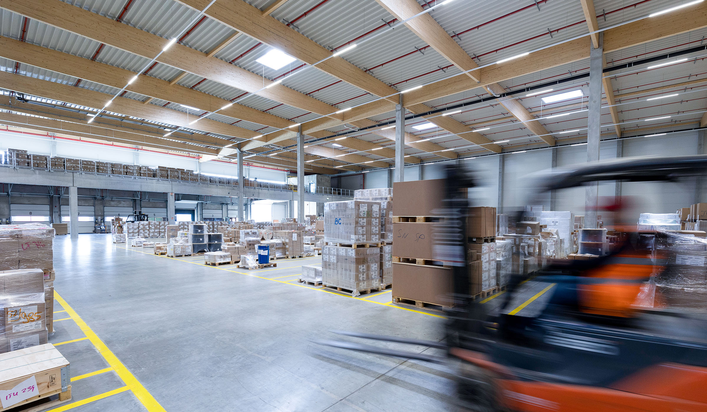 Lagerhalle mit Paletten und einem Gabelstapler, der schnell durch den Raum fährt. Helle Beleuchtung und Holzdecke sichtbar.