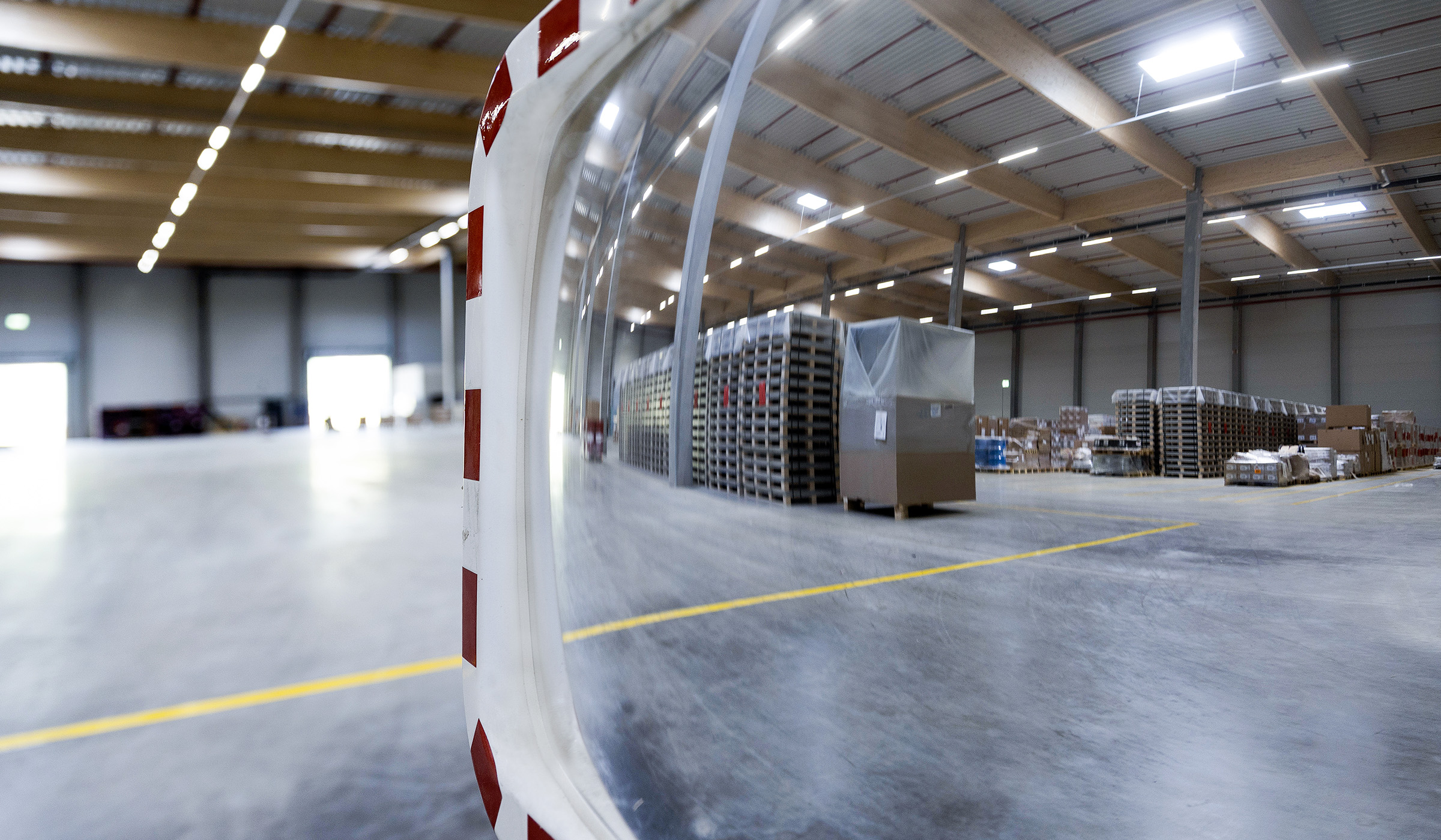 Reflexion in einem Sicherheits spiegel, mit einem großen, gut beleuchteten Lager im Hintergrund. Paletten und Verpackungen sind sichtbar.