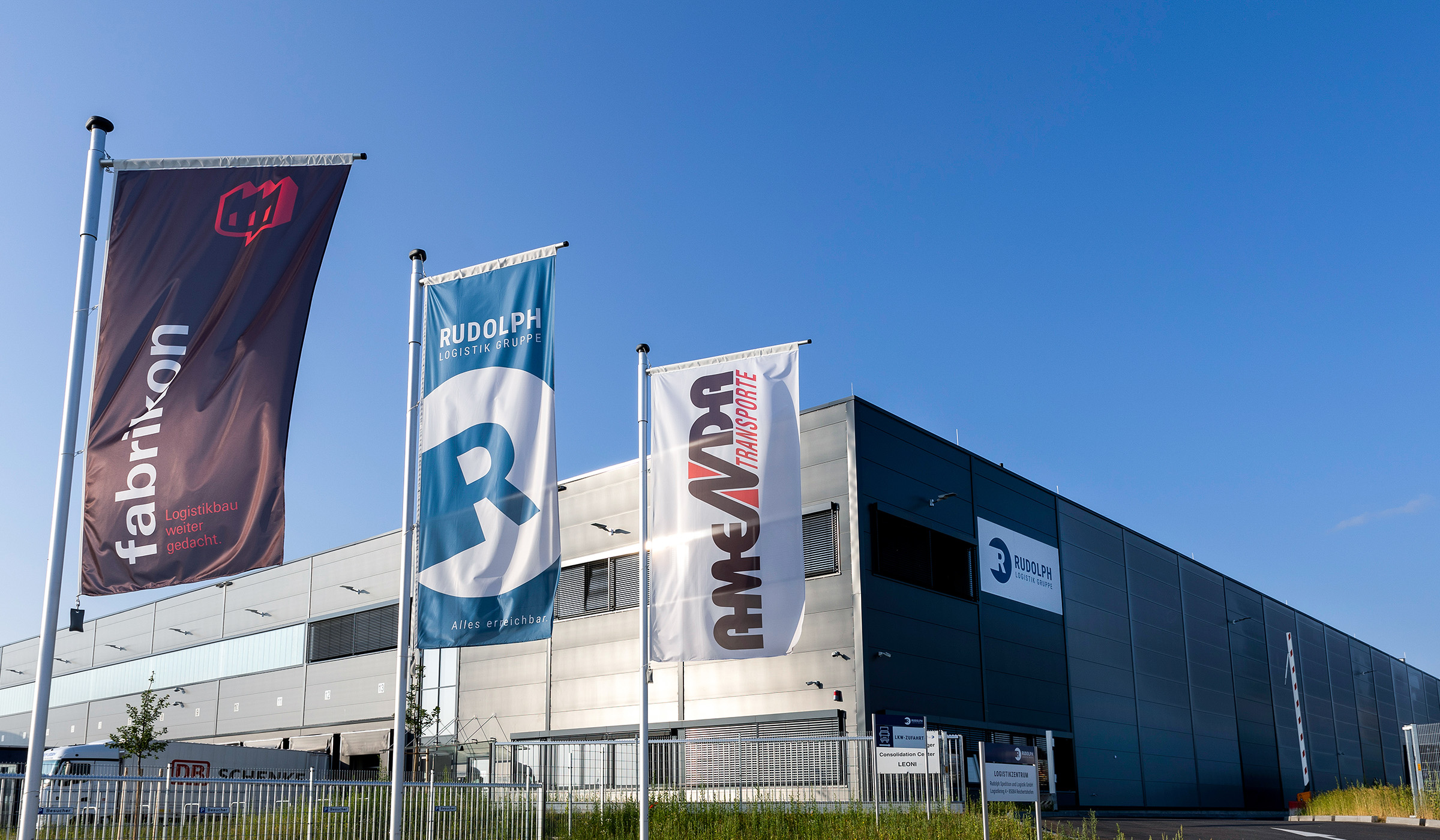 Drei Fahnen mit den Aufschriften "fabrikon", "RUDOLPH" und "AMEPARA Transporte" vor einem modernen Lagergebäude an blauen Himmel.