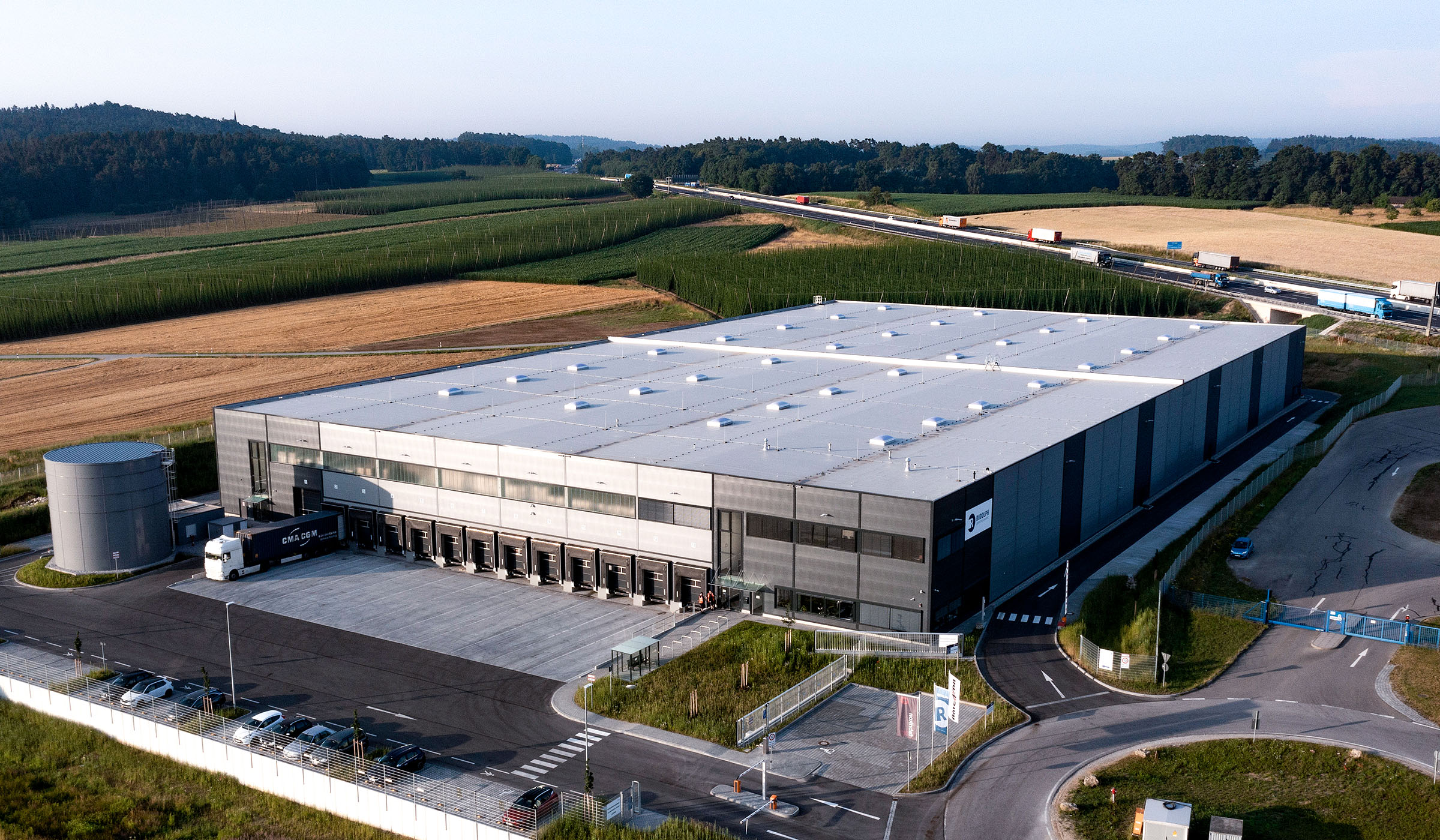 Luftaufnahme eines modernen Lagers mit Ladefläche, umgeben von landwirtschaftlichen Feldern und Fahrzeugen auf einer Straße.