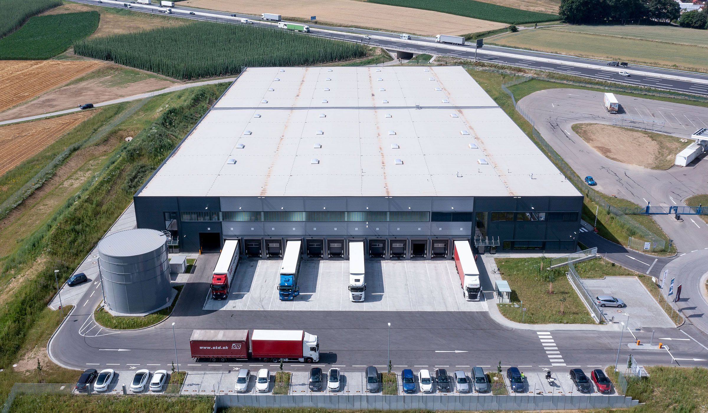 Luftaufnahme eines modernen Logistikgebäudes mit Lkw, Parkplätzen und umliegenden Feldern.