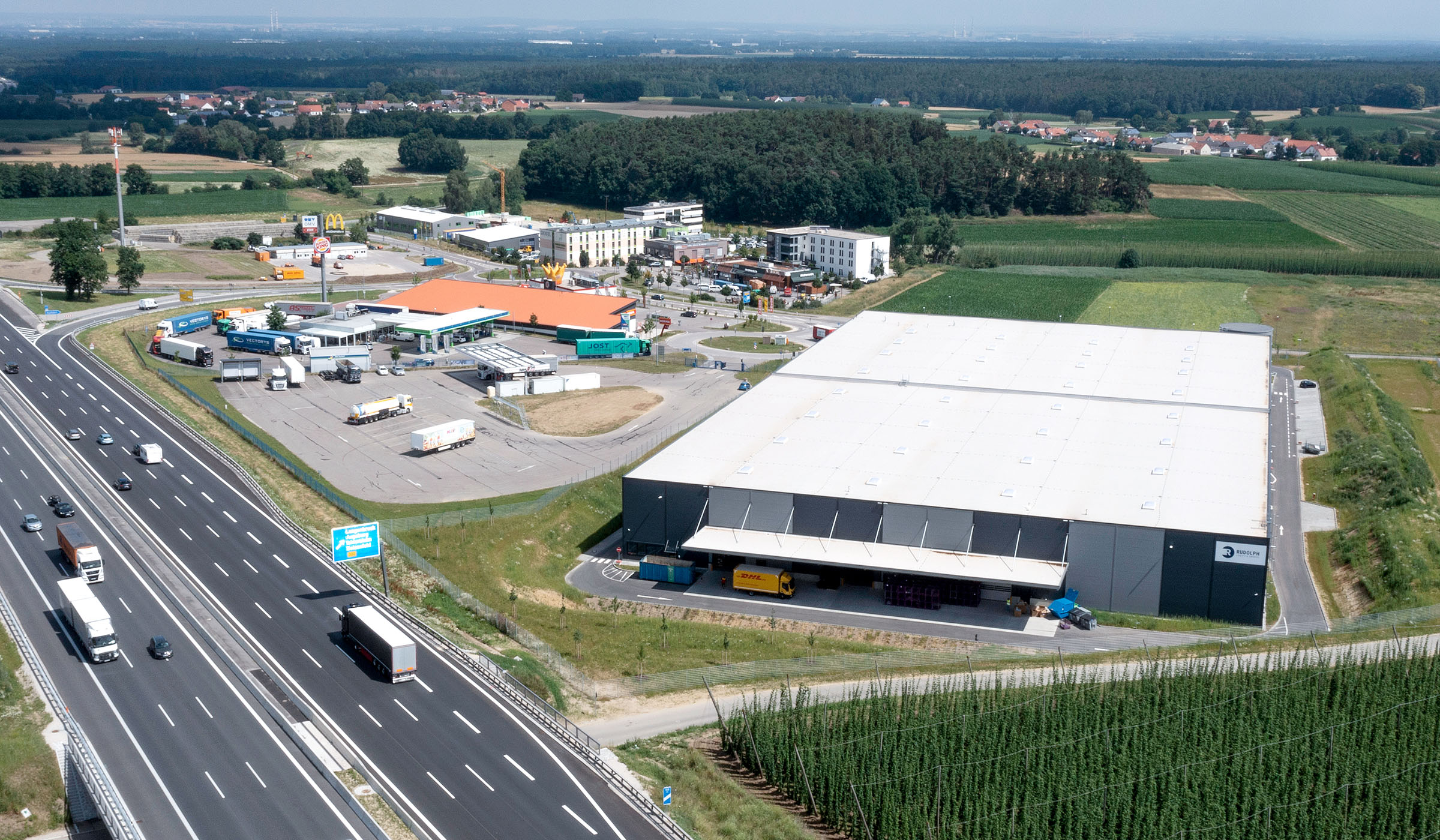 Luftaufnahme eines Logistikzentrums neben der Autobahn, umgeben von Feldern und einem kleinen Handelsgebiet.