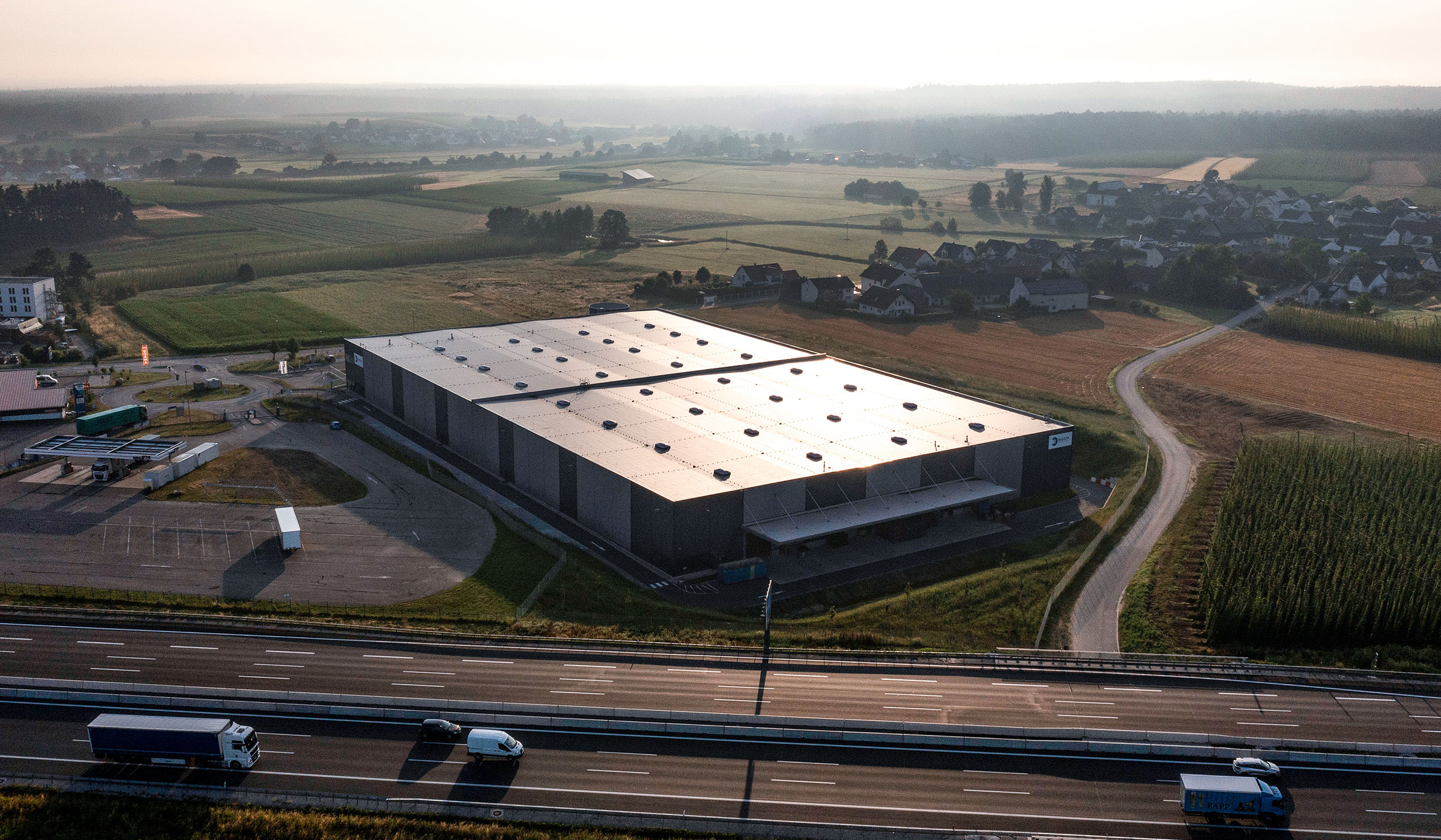 Ein modernes Lagergebäude mit Solarpanelen, umgeben von Feldern und einem sichtbaren Highway im Hintergrund.