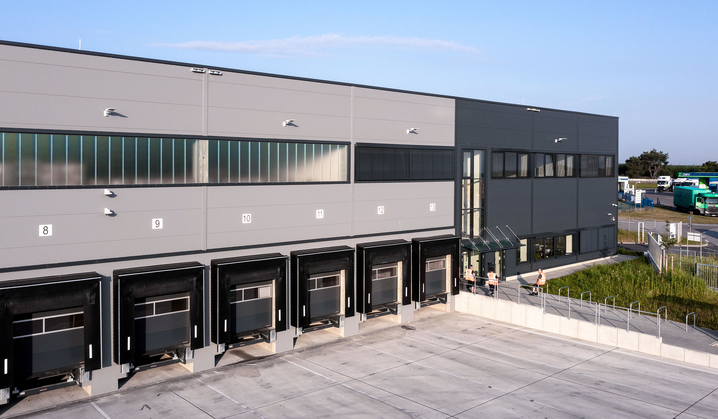 Moderne Lagerhalle mit Lkw-Ladebuchten und großen Fenstern, umgeben von Parkplatz und grünem Bereich.