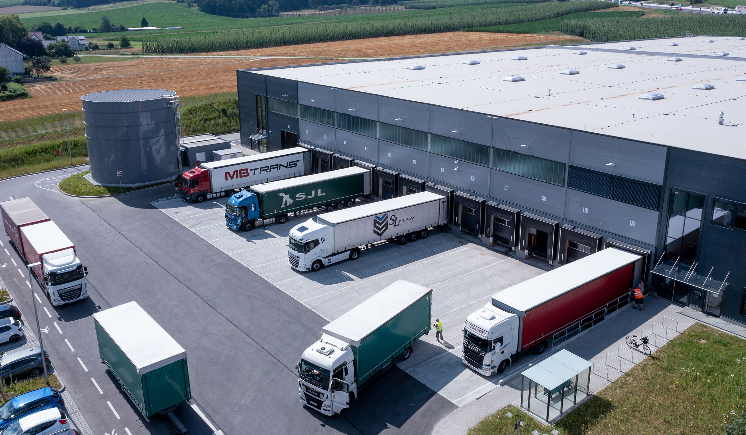 Luftaufnahme eines Lagerhauses mit mehreren Lkw und Ladebereichen, umgeben von Feldern und einem Wassertank.