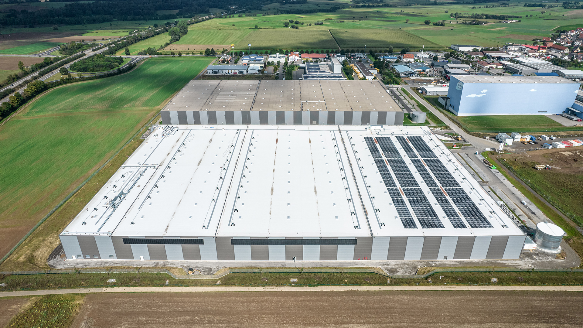 Luftaufnahme eines großen Lagergebäudes mit Solaranlagen auf dem Dach, umgeben von Feldern und anderen Gebäuden.