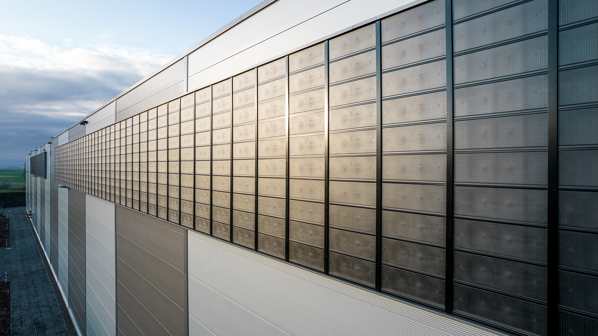 Große moderne Gebäudeoberfläche mit Fensterreihen und reflektierenden Materialien, umgeben von freiem Gelände und wolkigem Himmel.