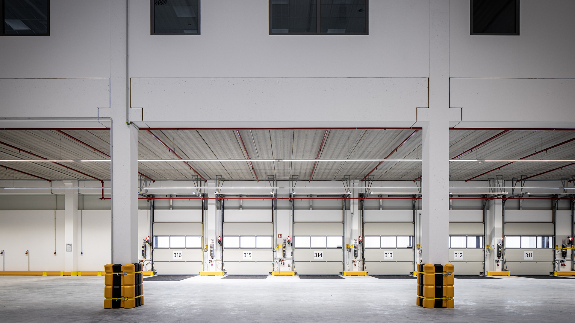 Lagerhalle mit mehreren Lkw-Ladeflächen, gelbe Schutzvorrichtungen und helle Decke. Grosse Fenster lassen Tageslicht herein.