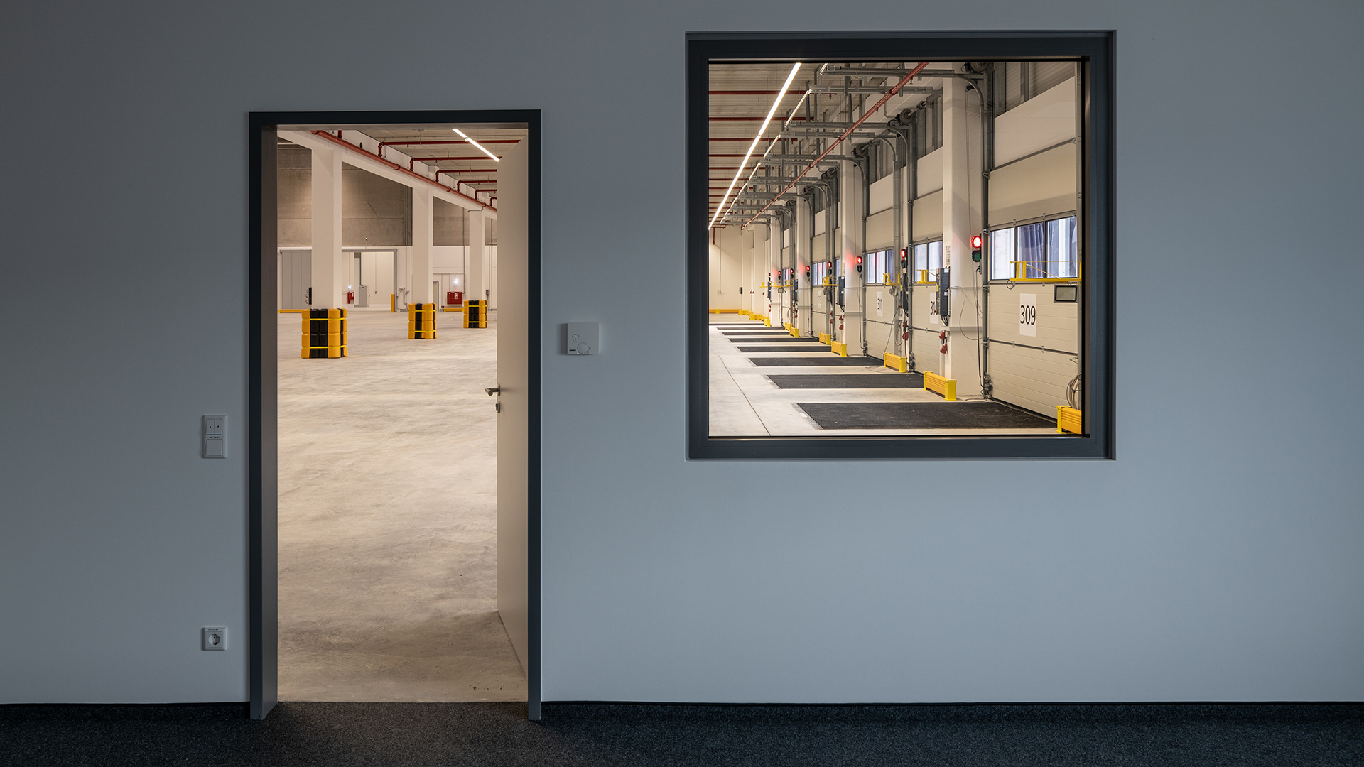 Blick durch eine Tür auf einen großen, modernen Lagerbereich mit Rolltoren und gelben Pylonen.