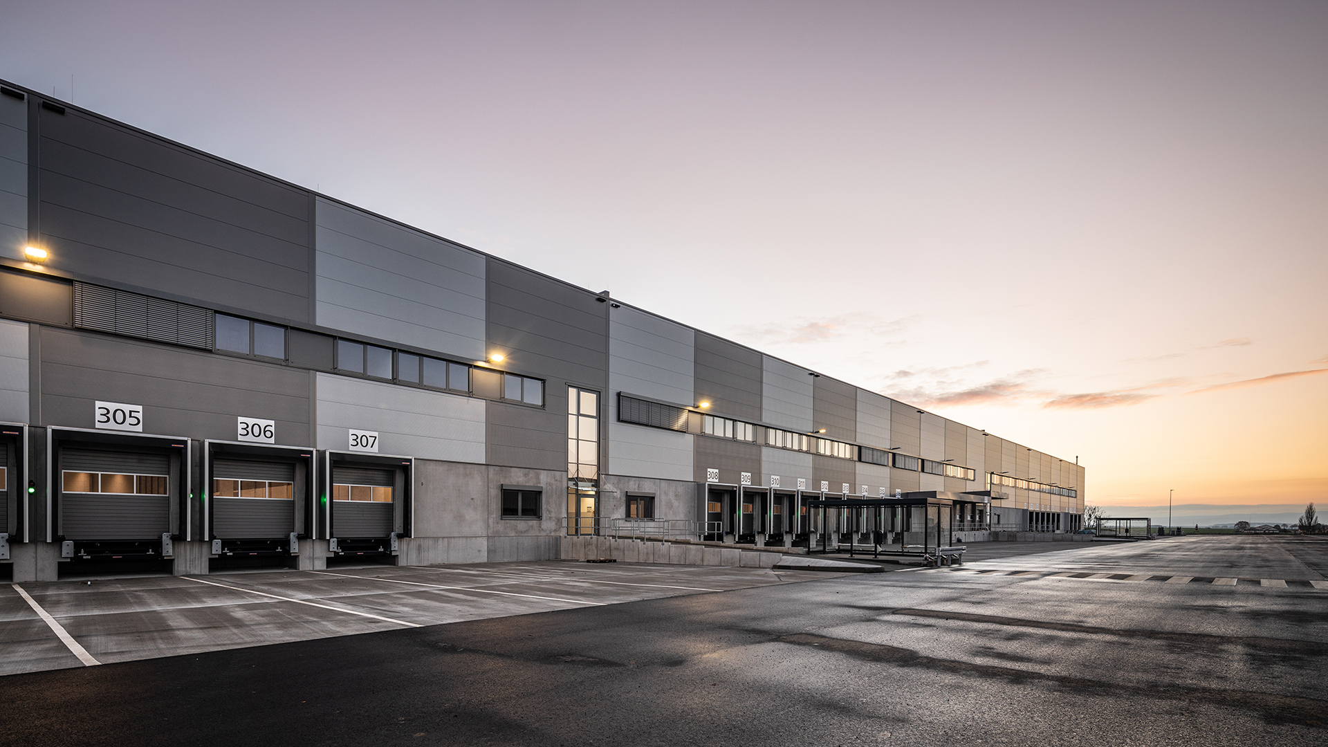 Moderne Lagerhalle bei Sonnenaufgang mit Laderampen und Parkplätzen im Vordergrund.