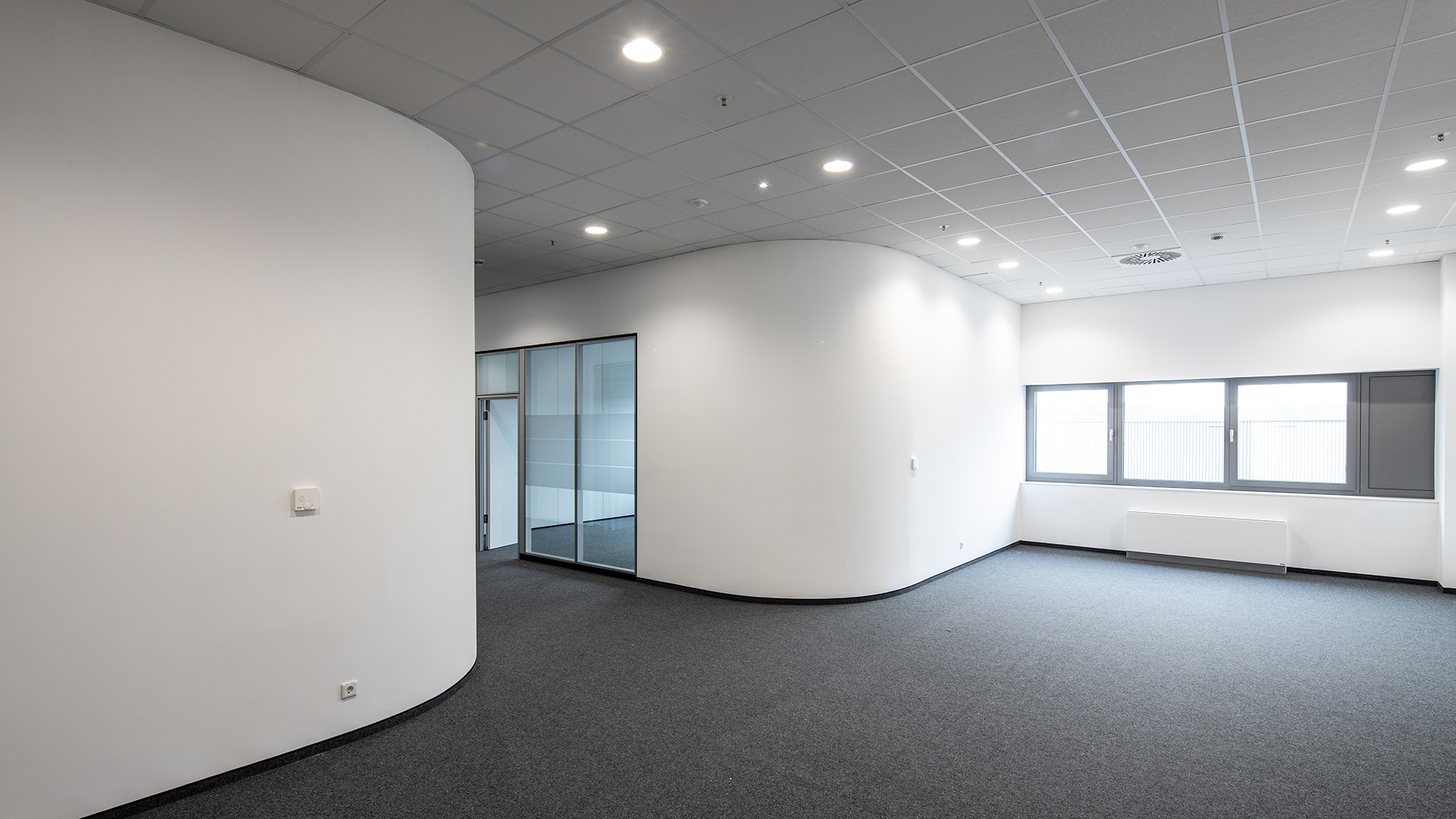 Minimalistisches Büro mit hellen Wänden, grauem Teppichboden und Fensterfront. Ruhe und offene Raumgestaltung sind vorhanden.