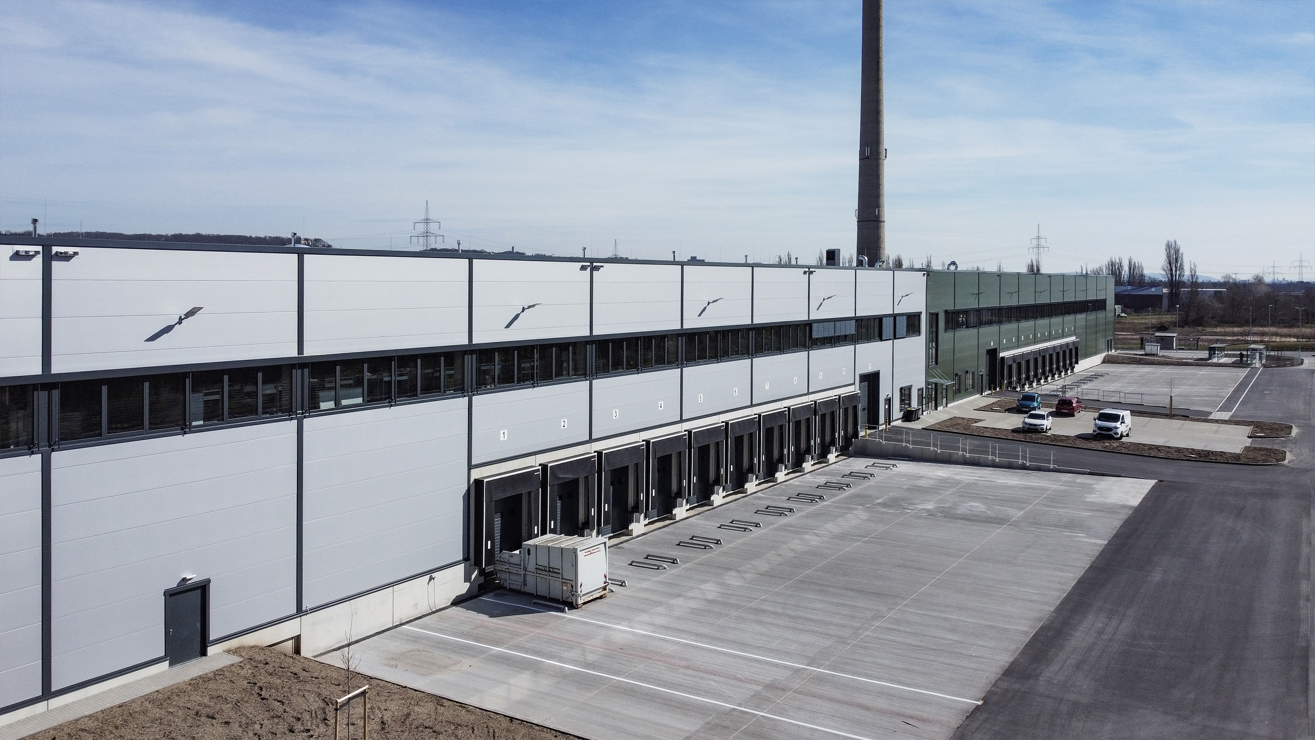 Moderne Lagerhalle mit Verladeflächen, geparkten Autos und einem hohen Schornstein im Hintergrund. Klarer blauer Himmel.
