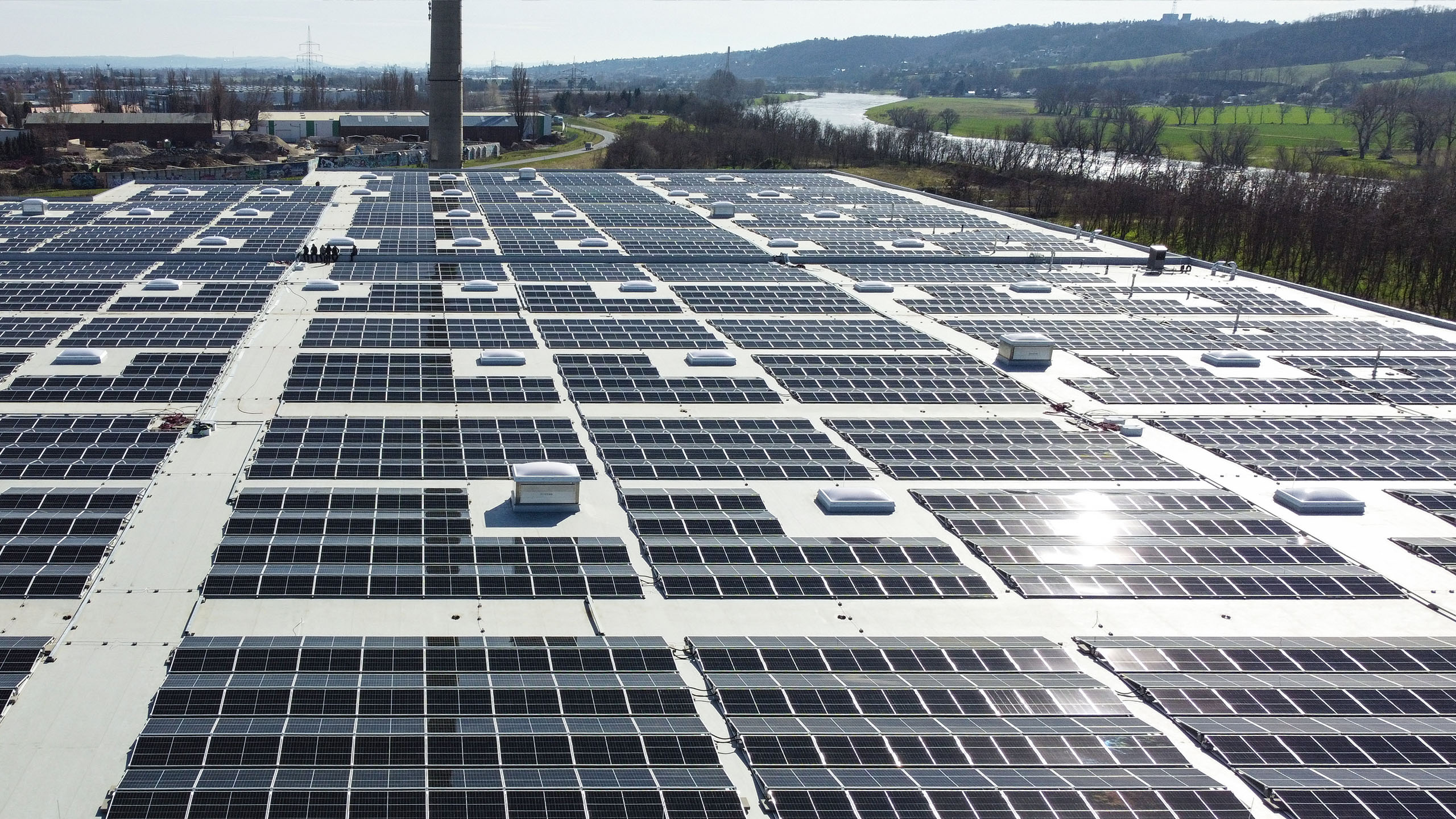 Photovoltaikanlage auf einem Flachdach mit mehreren Solarmodulen und Lichtkuppeln, umgeben von der Landschaft.