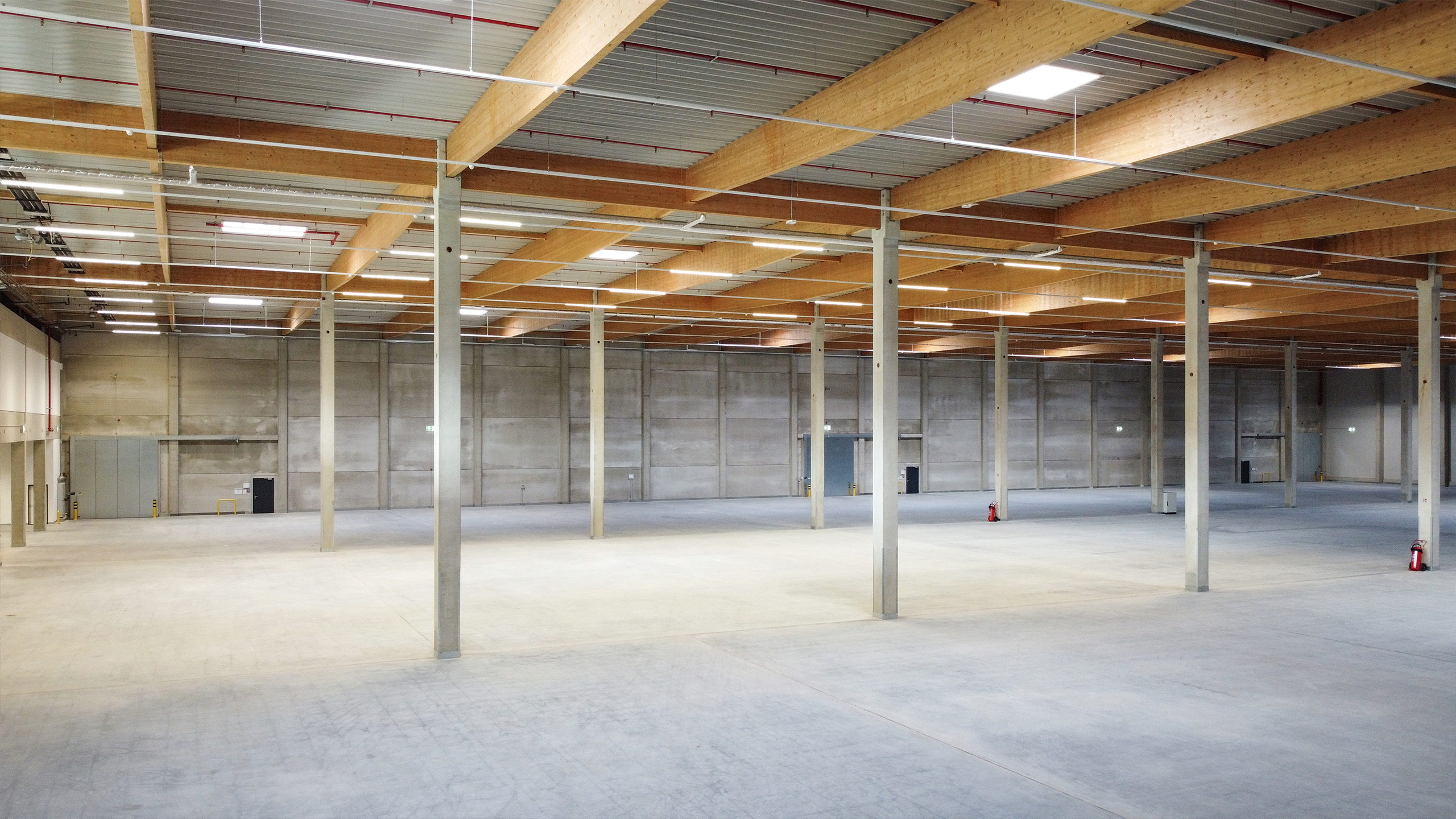 Weiträumiger, leerer Lagerraum mit hohen Decken, sichtbaren Holz- und Betonstrukturen sowie minimaler Ausstattung.