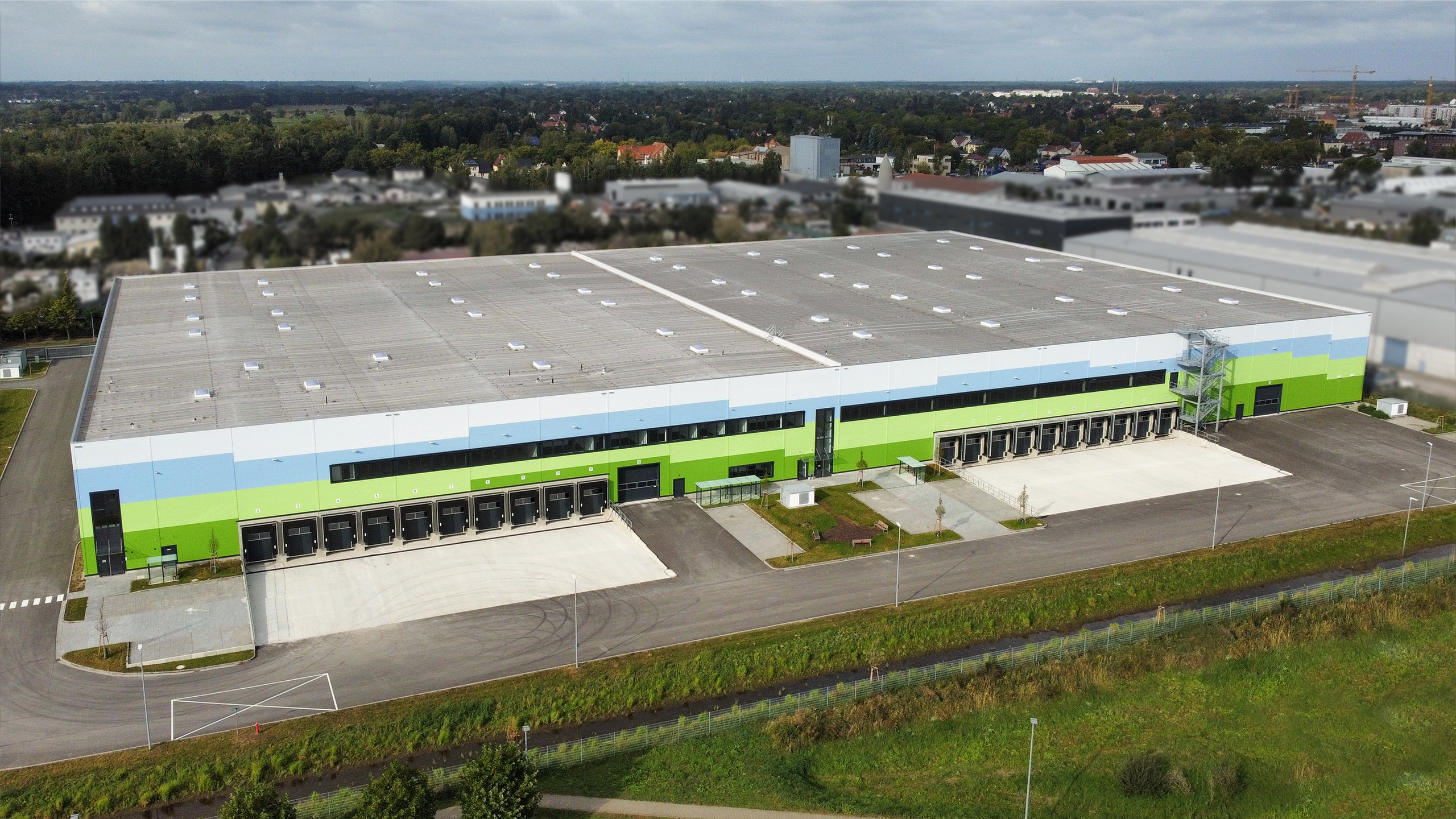 Ein modernes, mehrstöckiges Lagergebäude mit grüner und blauer Fassade sowie Lkw-Rampen und Parkplatz.